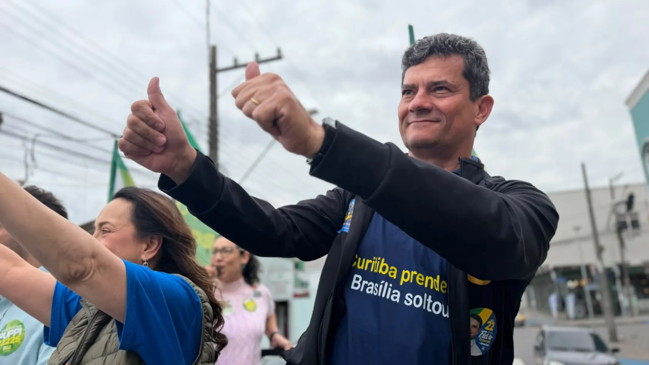 Fotografia em plano médio de ângulo contra-plongée (de baixo para cima) do senador Sergio Moro participando de ato público ou caminhada de campanha eleitoral em rua urbana. Em primeiro plano, à direita, destaca-se Sergio Moro, homem de pele clara e cabelos castanho-escuros curtos ligeiramente grisalhos. Enquadrado do peito para cima e levemente inclinado para a esquerda com um sorriso contido, ele veste uma jaqueta preta aberta sobre camiseta azul-marinho com a frase impressa "Curitiba prende, Brasília soltou". Ele está com os dois braços elevados para a frente fazendo o gesto de "positivo" com os polegares levantados. Na altura do peito, na jaqueta, há um adesivo de campanha político-eleitoral. No plano intermediário, à esquerda, veem-se apoiadores e correligionários com os braços erguidos vestindo roupas azuis e verdes com adesivos de campanha. Ao fundo, sob céu nublado de dia claro, observa-se a paisagem de uma via pública urbana com fiação elétrica, postes e fachadas comerciais.