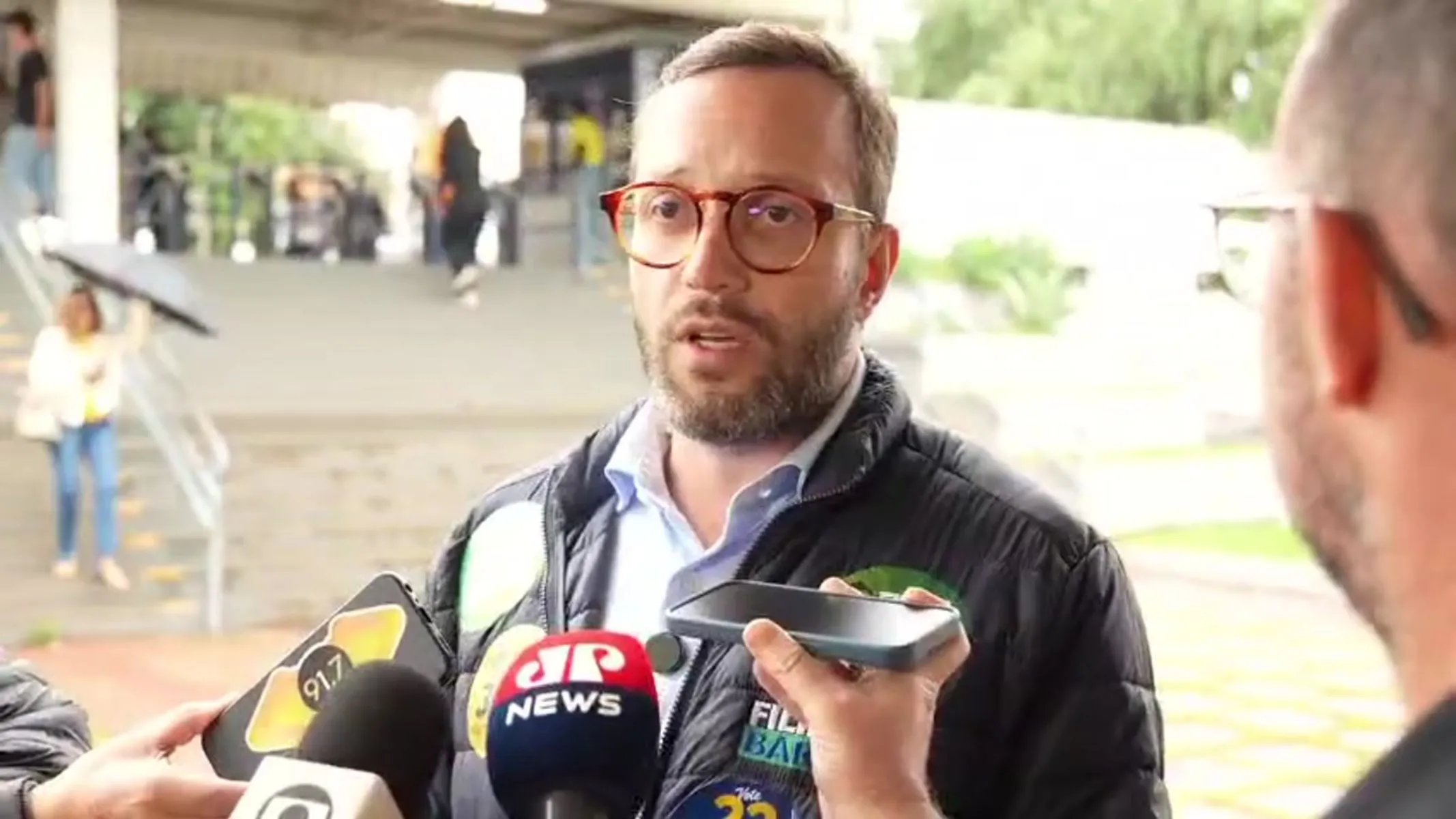 Fotografia em plano médio do deputado estadual Denian Couto concedendo entrevista a repórteres em ambiente externo coberto. Em primeiro plano, ligeiramente deslocado para a esquerda, destaca-se Denian Couto, homem de pele clara, cabelos castanho-escuros curtos, barba e bigode aparados e óculos de armação de acetato marrom-tartaruga. Ele veste uma jaqueta preta acolchoada tipo puffer sobre camisa azul-clara. Na altura do peito de sua jaqueta, há adesivos de campanha política. O parlamentar está voltado para a direita com a boca ligeiramente aberta enquanto fala. À sua frente e ao seu redor, veem-se as mãos e os braços de repórteres segurando microfones (incluindo um com a canopla vermelha da "JP NEWS") e smartphones para gravar a declaração. No canto inferior direito, vê-se de costas a parte posterior da cabeça e a orelha de um dos entrevistadores. Ao fundo, observa-se a área externa com escadaria de concreto, pilares e pedestres circulando sob iluminação natural.