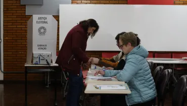 Fotografia em plano médio do interior de uma seção eleitoral durante o processo de identificação e votação de eleitores. Em primeiro plano, do centro para a direita, veem-se eleitores e mesários posicionados junto a uma mesa de atendimento. Em destaque no canto direito, uma pessoa de cabelos curtos loiros e óculos veste uma jaqueta acolchoada puffer azul-clara e está sentada à mesa segurando uma caneta. Ao seu lado, no centro da imagem, uma mulher de pele clara, cabelos escuros na altura dos ombros e casaco vinho escuro está de pé, inclinando-se ligeiramente sobre a mesa para realizar a identificação biométrica ou assinar o caderno de votação. No plano intermediário, à esquerda, sobre uma mesa individual de madeira, posiciona-se a cabina de votação de papelão branco com a urna eletrônica acoplada, exibindo as inscrições "JUSTIÇA ELEITORAL", o Brasão da República e "CABINA DE VOTAÇÃO". Ao fundo, observa-se a parede da sala com revestimento de tijolos aparentes à esquerda, quadros brancos escolares e cadeiras alinhadas sob iluminação artificial uniforme de ambiente interno.