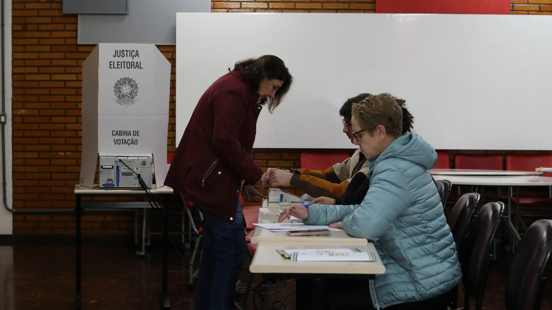 Fotografia em plano médio do interior de uma seção eleitoral durante o processo de identificação e votação de eleitores. Em primeiro plano, do centro para a direita, veem-se eleitores e mesários posicionados junto a uma mesa de atendimento. Em destaque no canto direito, uma pessoa de cabelos curtos loiros e óculos veste uma jaqueta acolchoada puffer azul-clara e está sentada à mesa segurando uma caneta. Ao seu lado, no centro da imagem, uma mulher de pele clara, cabelos escuros na altura dos ombros e casaco vinho escuro está de pé, inclinando-se ligeiramente sobre a mesa para realizar a identificação biométrica ou assinar o caderno de votação. No plano intermediário, à esquerda, sobre uma mesa individual de madeira, posiciona-se a cabina de votação de papelão branco com a urna eletrônica acoplada, exibindo as inscrições "JUSTIÇA ELEITORAL", o Brasão da República e "CABINA DE VOTAÇÃO". Ao fundo, observa-se a parede da sala com revestimento de tijolos aparentes à esquerda, quadros brancos escolares e cadeiras alinhadas sob iluminação artificial uniforme de ambiente interno.