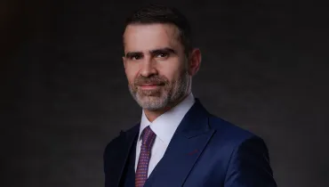 Fotografia em primeiro plano do tipo retrato corporativo e institucional de um homem em estúdio.Em primeiro plano, centralizado na imagem, destaca-se um homem de pele clara, cabelos castanho-escuros curtos e barba grisalha bem aparada. Enquadrado do peito para cima e voltado de três quartos para a esquerda, ele olha diretamente para a câmera com um sorriso leve e expressão serena. Ele veste traje formal composto por terno azul-marinho de abotoamento elegante, colete azul-marinho do mesmo tecido sob o paletó, camisa social branca de gola estruturada e gravata em tom grená ou vinho com pequena estampa geométrica repetitiva. Ao fundo, o cenário é composto por um fundo infinito fotográfico cinza-escuro neutro e texturizado. A iluminação de estúdio é lateral e bem direcionada, criando um contraste suave entre luz e sombra no rosto do retratado e destacando os detalhes do traje.