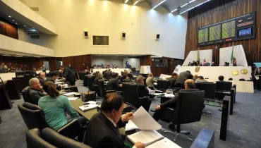 Fotografia em plano geral amplo registrada de um ponto elevado (plongée) da galeria de visitantes do Plenário da Assembleia Legislativa do Estado do Paraná (ALEP). Em primeiro plano, de costas para a câmera, observa-se o público e populares lotando a arquibancada em anfiteatro.