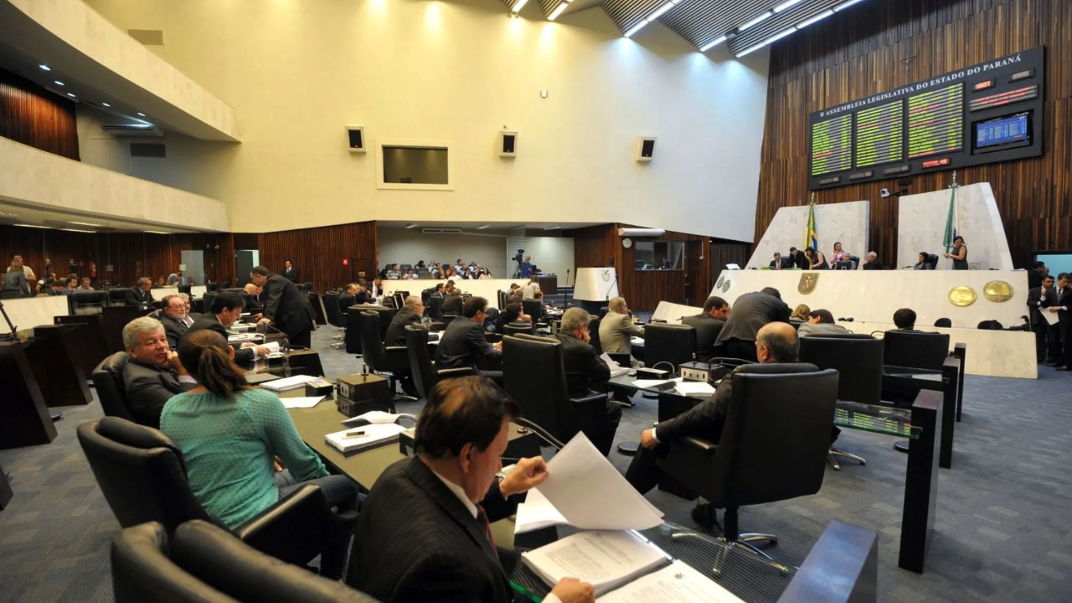 Fotografia em plano geral amplo registrada de um ponto elevado (plongée) da galeria de visitantes do Plenário da Assembleia Legislativa do Estado do Paraná (ALEP). Em primeiro plano, de costas para a câmera, observa-se o público e populares lotando a arquibancada em anfiteatro.