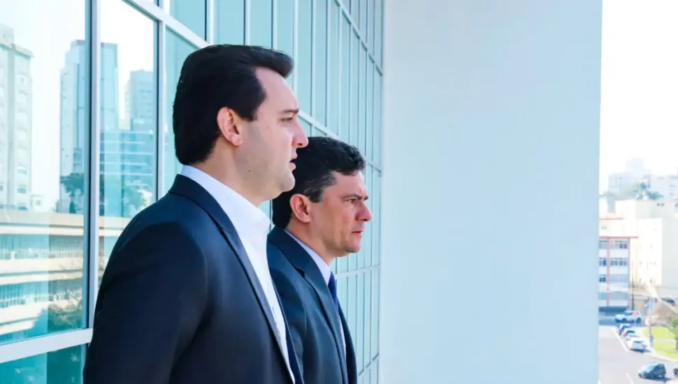 Fotografia em plano médio e enquadramento em perfil lateral do governador do Paraná, Ratinho Junior, e do senador Sergio Moro em ambiente institucional moderno. Em primeiro plano, à esquerda, destaca-se Ratinho Junior, homem de pele clara e cabelos castanho-escuros curtos penteados para trás. Visto de perfil e enquadrado do peito para cima, ele veste paletó preto sobre camisa social branca sem gravata. Logo ao seu lado, ligeiramente atrás e no plano intermediário, posiciona-se Sergio Moro, homem de pele clara e cabelos curtos escuros, vestindo terno escuro e gravata azul-marinho. Ambos olham atentamente na mesma direção, voltados para a direita da imagem. No plano intermediário e ao fundo, os dois homens estão posicionados junto a uma fachada de estrutura moderna composta por grandes painéis e janelas de vidro espelhado que refletem o ambiente urbano exterior. À direita, abre-se a vista em perspectiva para uma área externa e edifícios sob luz natural do dia clara e uniforme.