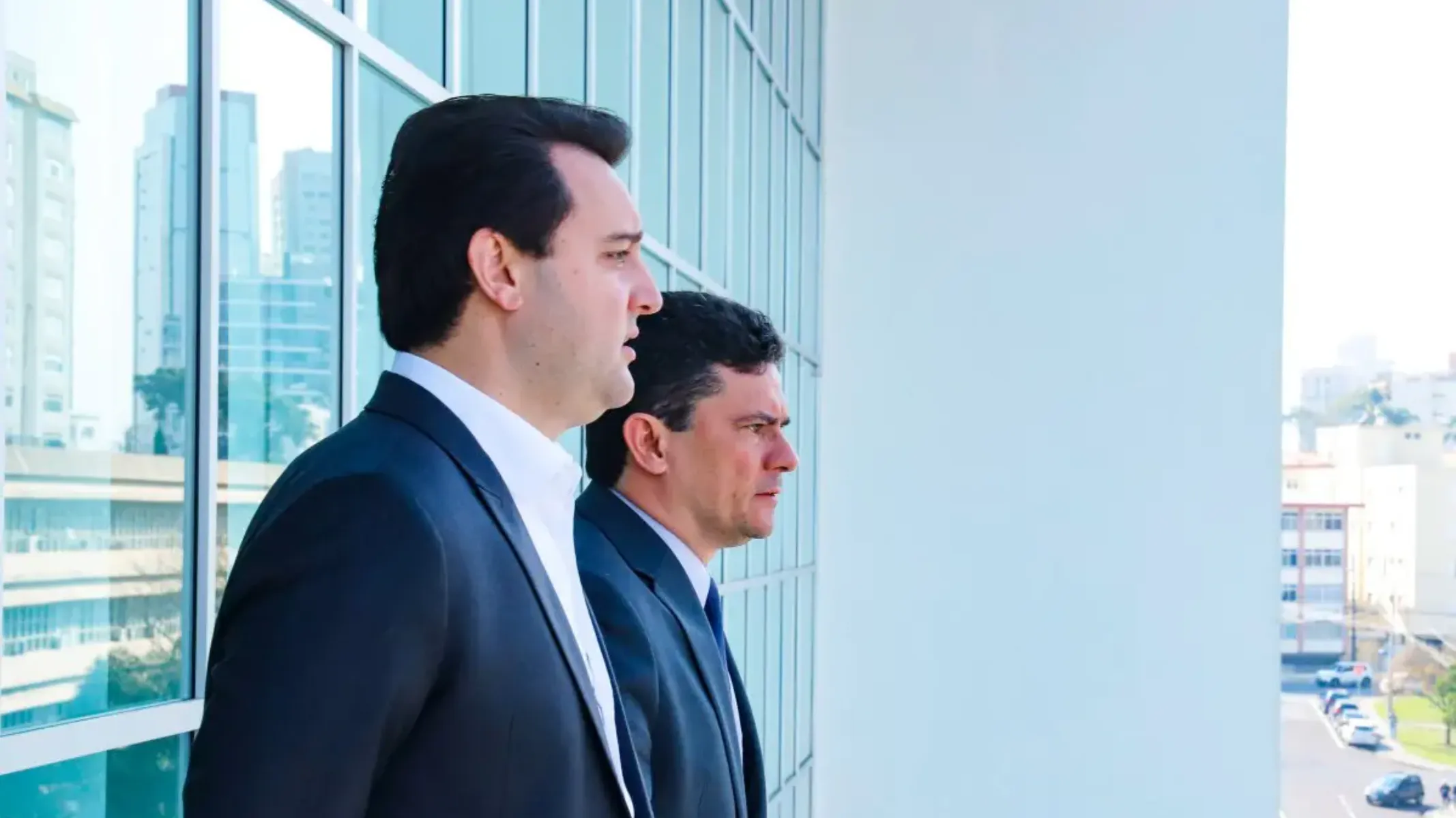 Fotografia em plano médio e enquadramento em perfil lateral do governador do Paraná, Ratinho Junior, e do senador Sergio Moro em ambiente institucional moderno. Em primeiro plano, à esquerda, destaca-se Ratinho Junior, homem de pele clara e cabelos castanho-escuros curtos penteados para trás. Visto de perfil e enquadrado do peito para cima, ele veste paletó preto sobre camisa social branca sem gravata. Logo ao seu lado, ligeiramente atrás e no plano intermediário, posiciona-se Sergio Moro, homem de pele clara e cabelos curtos escuros, vestindo terno escuro e gravata azul-marinho. Ambos olham atentamente na mesma direção, voltados para a direita da imagem. No plano intermediário e ao fundo, os dois homens estão posicionados junto a uma fachada de estrutura moderna composta por grandes painéis e janelas de vidro espelhado que refletem o ambiente urbano exterior. À direita, abre-se a vista em perspectiva para uma área externa e edifícios sob luz natural do dia clara e uniforme.