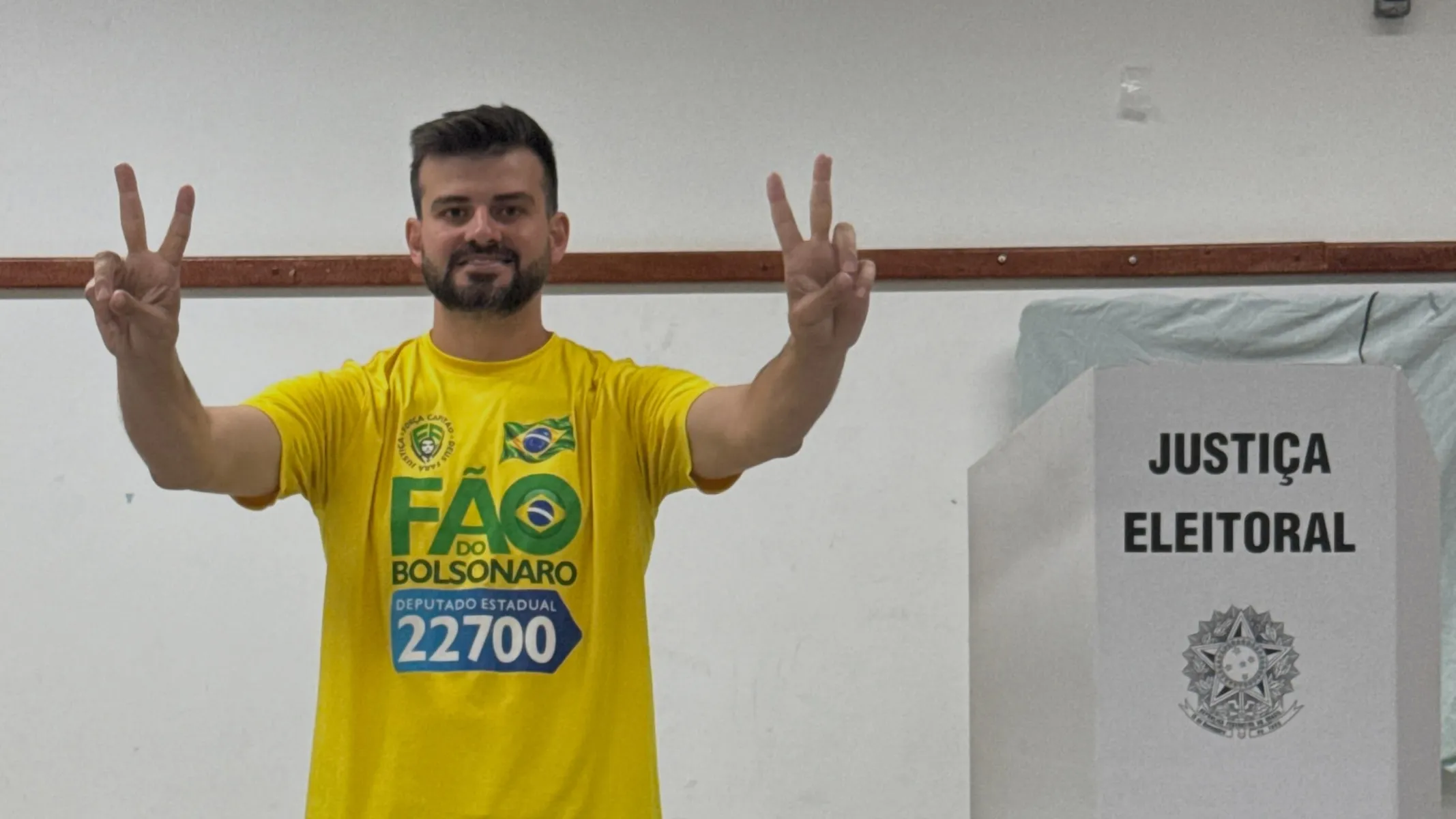 Fotografia em plano médio de um candidato a deputado estadual em seção eleitoral no momento da votação. Em primeiro plano, do centro para o lado esquerdo da imagem, destaca-se um homem de pele clara, cabelos castanho-escuros curtos e barba aparada, sorrindo diretamente para a câmera. Enquadrado da cintura para cima, ele veste uma camiseta amarela de campanha eleitoral estampada com o nome "FÃO DO BOLSONARO", o cargo "DEPUTADO ESTADUAL" e o número "22700", além da bandeira do Brasil no peito. Ele está com os dois braços erguidos fazendo o sinal de "V" de vitória com os dedos indicador e médio de ambas as mãos. No plano intermediário, à direita, posiciona-se uma cabina de votação de papelão cinza-claro da Justiça Eleitoral, exibindo o texto "JUSTIÇA ELEITORAL" em caixa-alta preta acima do Brasão da República. Ao fundo, observa-se uma parede lisa de tom claro com um rodapé de madeira sob iluminação artificial interna e uniforme.