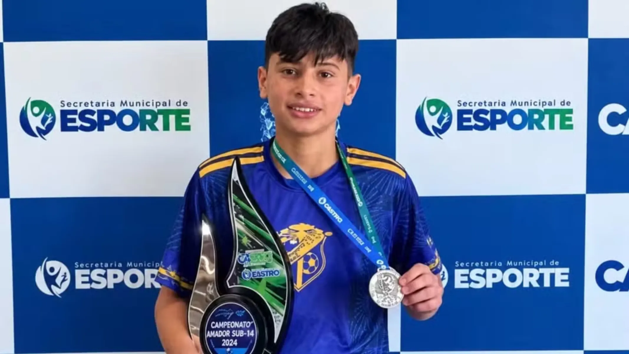 Fotografia de um menino vestindo uma camiseta azul. Ele segura uma medalha e um troféu.