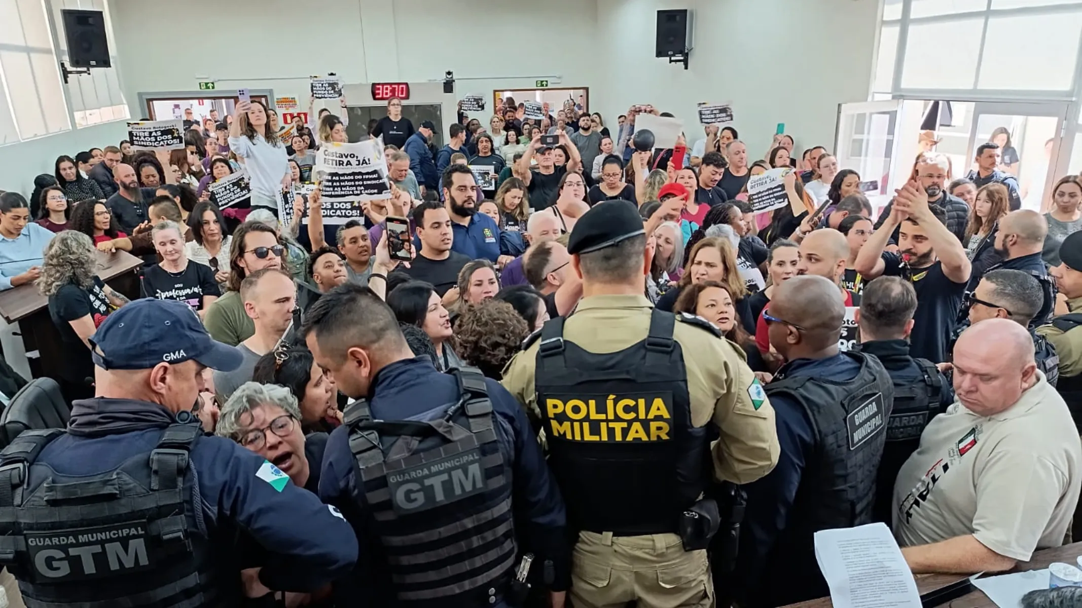Fotografia em plano geral de uma sessão pública marcada por manifestação, tumulto e presença de forças de segurança em auditório institucional. Em primeiro plano, de costas para a câmera, posicionam-se agentes de segurança pública atuando como cordão de contenção. No centro, destaca-se um policial militar vestindo farda caqui com colete tático preto no qual se lê "POLÍCIA MILITAR" em letras amarelas em destaque. À sua esquerda e à sua direita, agentes da Guarda Municipal e do Grupo Tático Municipal (GTM) vestem uniformes azuis-escuros e coletes táticos pretos com as inscrições "GUARDA MUNICIPAL" e "GTM" em cinza e branco nas costas. No canto inferior direito, um homem calvo de pele clara veste camiseta polo bege e segura documentos sobre uma mesa. No plano intermediário, diante dos agentes de segurança, uma multidão de manifestantes, servidores e populares aglomera-se em tom de protesto. Várias pessoas gesticulam, gritam, filmam com celulares ou erguem cartazes e faixas com reivindicações e críticas. Ao fundo, o salão do auditório apresenta paredes claras, grandes janelas de vidro e caixas de som fixadas no alto, sob iluminação artificial uniforme do ambiente interno.