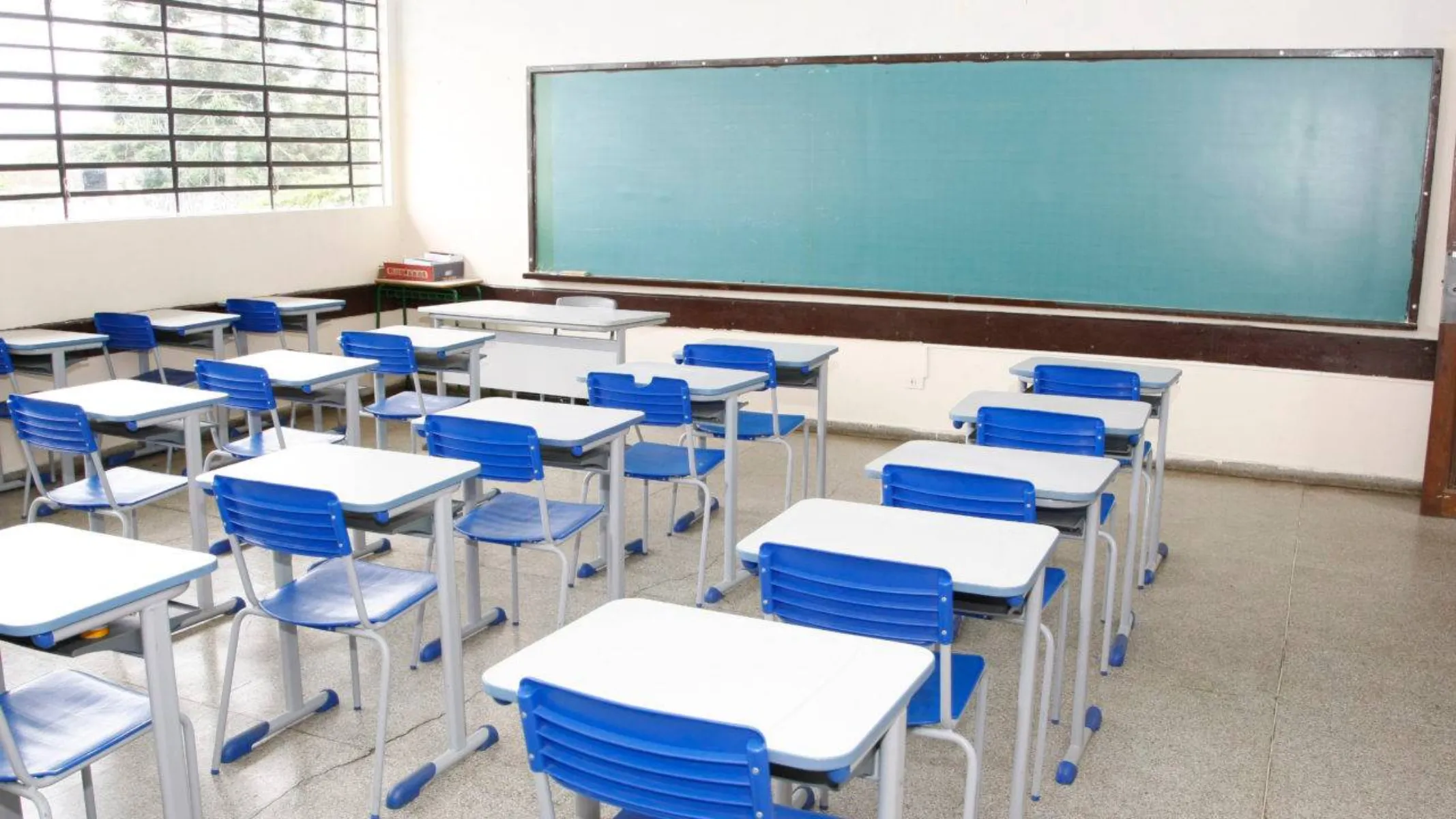 Fotografia em plano geral do interior de uma sala de aula escolar vazia e organizada. Em primeiro plano e no plano intermediário, observam-se várias fileiras paralelas de conjuntos escolares compostos por carteiras individuais com tampos claros de formato retangular e cadeiras de estrutura metálica cinza com assento e encosto de plástico azul-vivo, todas alinhadas e sem alunos. Ao fundo, na parede principal de tom claro, estende-se na horizontal um grande quadro-negro escolar de superfície verde-escura emoldurado por friso metálico. À esquerda, há uma ampla janela com grades e basculantes de vidro que permite a entrada de luz natural abundante no ambiente, iluminando o piso claro e polido da sala.
