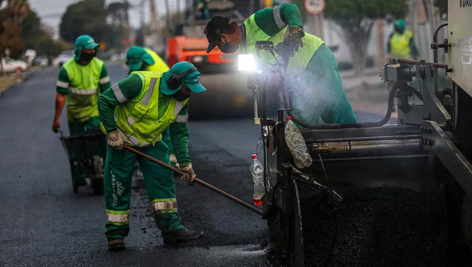Fotografia em plano médio de trabalhadores executando serviços de recapeamento asfáltico e obras de infraestrutura viária urbana sob luz natural. Em primeiro plano, do centro para a esquerda, destaca-se um operário curvado trabalhando no nivelamento da massa asfáltica com uma ferramenta manual de cabo longo. Ele veste uniforme de segurança composto por calça verde-escura com a inscrição "CARAVAGGIO" na perna, colete verde-limão refletivo sobre camisa de mangas compridas, boné árabe de proteção solar com aba estendida, protetor auricular e luvas. À sua direita, em primeiro plano, vê-se a lateral metálica de uma máquina pavimentadora de asfalto despejando a massa escura sobre o piso. No plano intermediário, um segundo trabalhador inclina-se sobre a estrutura da pavimentadora ajustando o maquinário sob uma luz de LED acesa, enquanto outro operário ao fundo conduz um carrinho de mão sobre o asfalto novo. Um rolo compressor alaranjado posiciona-se mais atrás no centro da via. Ao fundo, observa-se a perspectiva da rua em obras com árvores, veículos estacionados e placas de trânsito.