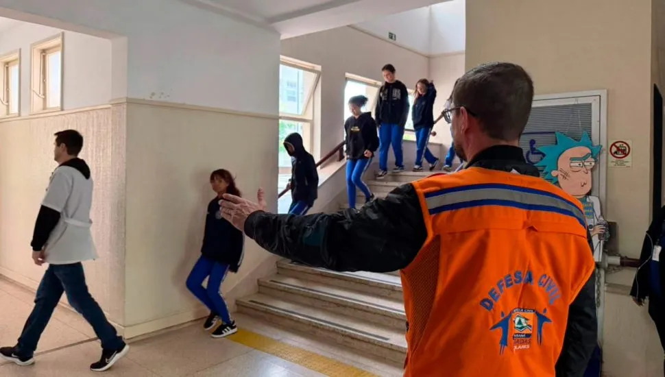 A imagem mostra um agente da Defesa Civil orientando a saída de alunos do prédio escolar.