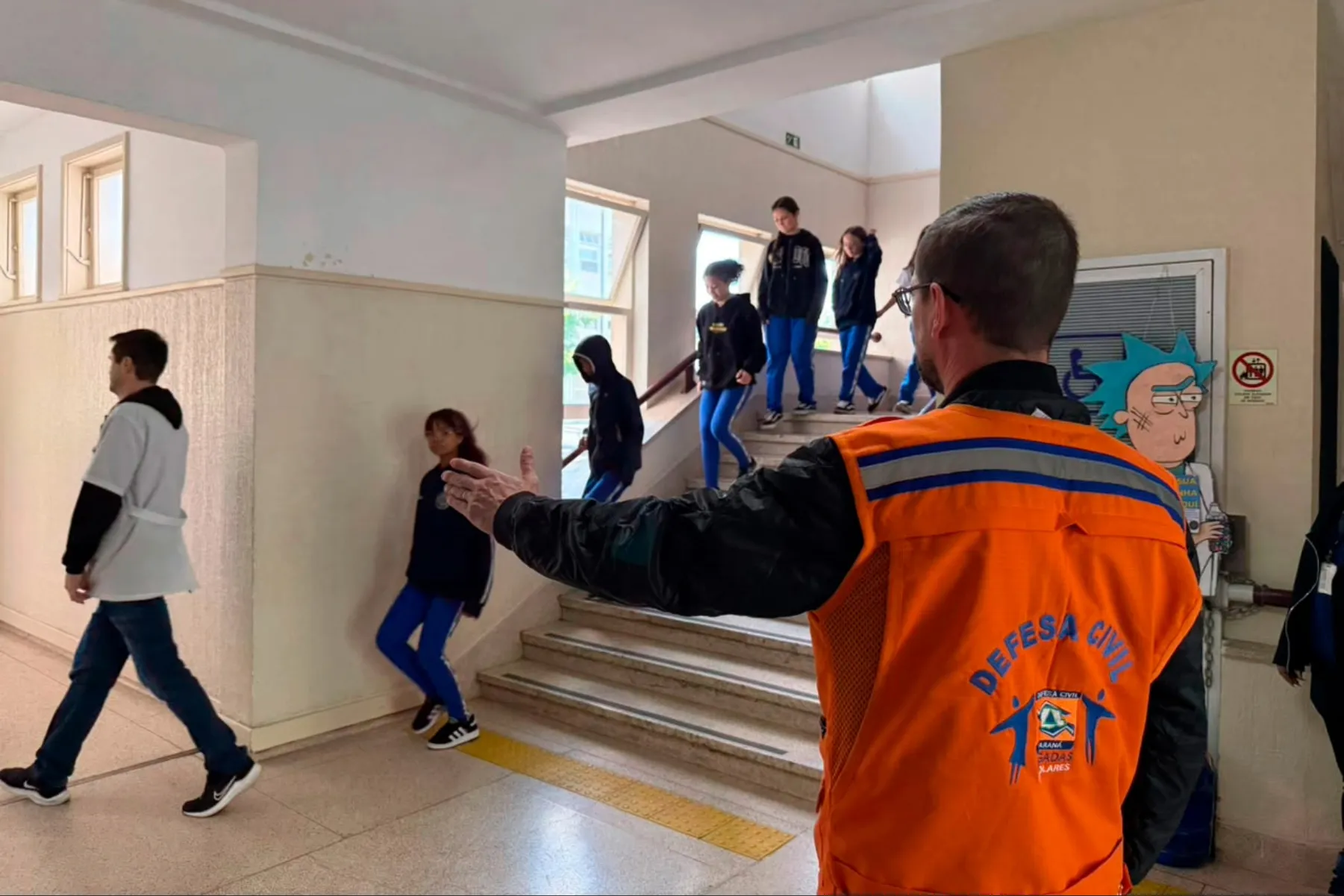 A imagem mostra um agente da Defesa Civil orientando a saída de alunos do prédio escolar.