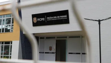 Fotografia em plano médio da fachada da Divisão de Homicídios e Proteção à Pessoa (DHPP) da Polícia Civil do Estado do Paraná (PCPR). Em primeiro plano, desfocados pela pouca profundidade de campo, veem-se os gradis e fios metálicos brancos de uma cerca ou portão que emolduram a cena. No plano intermediário, em foco, destaca-se a fachada principal do prédio, revestida por azulejos retangulares brancos. Acima da porta de entrada principal, sobressai uma placa retangular preta com o brasão da corporação à esquerda, seguido da sigla "PCPR" e do texto em letras brancas: "POLÍCIA CIVIL DO PARANÁ - DIVISÃO DE HOMICÍDIOS E PROTEÇÃO À PESSOA". Logo abaixo, a porta de acesso em alumínio branco exibe adesivos pretos horizontais com a sigla "PCPR" e o brasão policial centralizado. À direita, há um poste metálico escuro com luminárias de LED.
