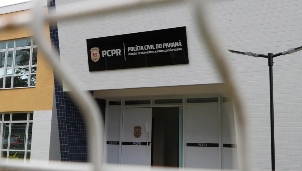 Fotografia em plano médio da fachada da Divisão de Homicídios e Proteção à Pessoa (DHPP) da Polícia Civil do Estado do Paraná (PCPR). Em primeiro plano, desfocados pela pouca profundidade de campo, veem-se os gradis e fios metálicos brancos de uma cerca ou portão que emolduram a cena. No plano intermediário, em foco, destaca-se a fachada principal do prédio, revestida por azulejos retangulares brancos. Acima da porta de entrada principal, sobressai uma placa retangular preta com o brasão da corporação à esquerda, seguido da sigla "PCPR" e do texto em letras brancas: "POLÍCIA CIVIL DO PARANÁ - DIVISÃO DE HOMICÍDIOS E PROTEÇÃO À PESSOA". Logo abaixo, a porta de acesso em alumínio branco exibe adesivos pretos horizontais com a sigla "PCPR" e o brasão policial centralizado. À direita, há um poste metálico escuro com luminárias de LED.