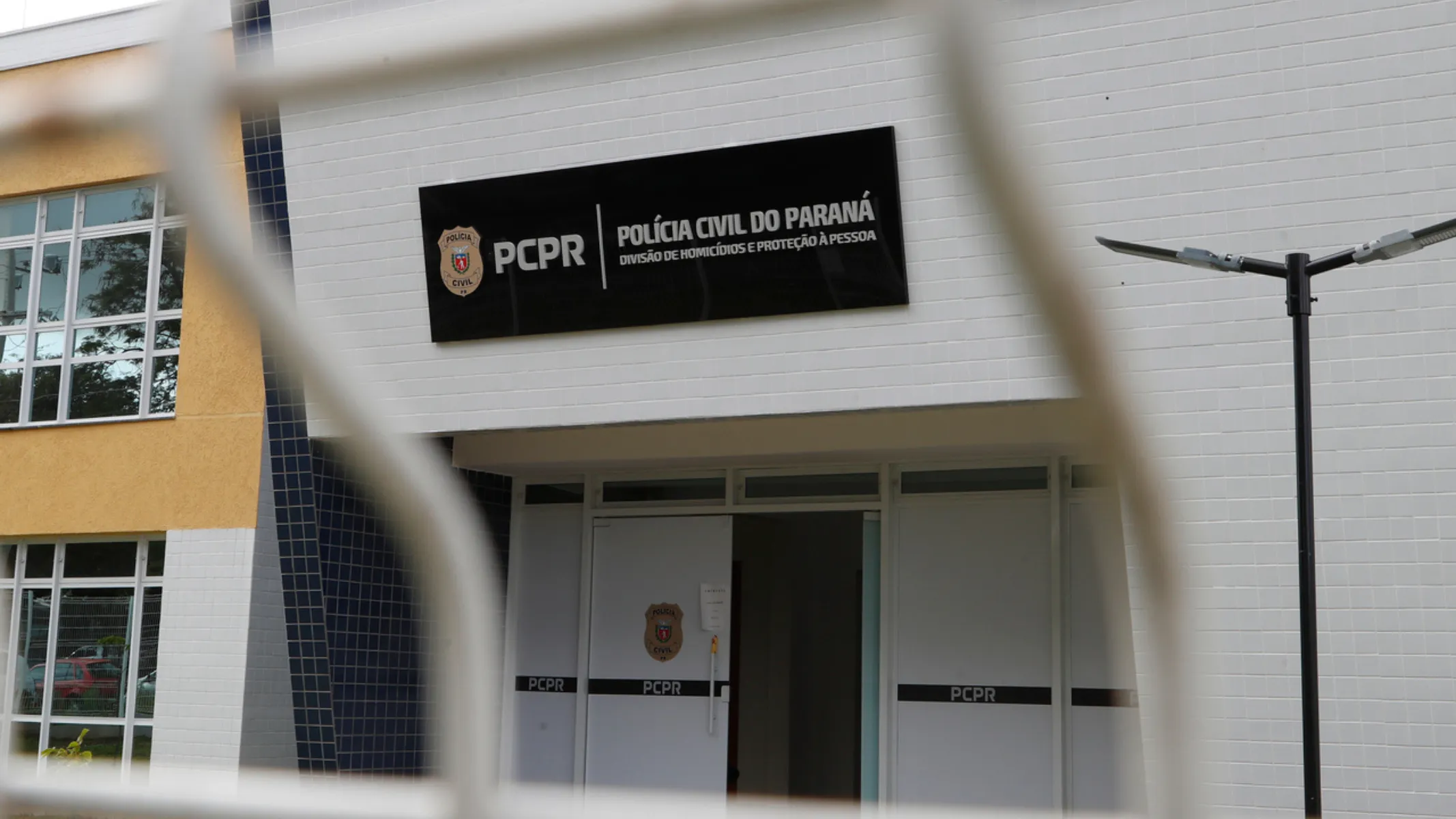 Fotografia em plano médio da fachada da Divisão de Homicídios e Proteção à Pessoa (DHPP) da Polícia Civil do Estado do Paraná (PCPR). Em primeiro plano, desfocados pela pouca profundidade de campo, veem-se os gradis e fios metálicos brancos de uma cerca ou portão que emolduram a cena. No plano intermediário, em foco, destaca-se a fachada principal do prédio, revestida por azulejos retangulares brancos. Acima da porta de entrada principal, sobressai uma placa retangular preta com o brasão da corporação à esquerda, seguido da sigla "PCPR" e do texto em letras brancas: "POLÍCIA CIVIL DO PARANÁ - DIVISÃO DE HOMICÍDIOS E PROTEÇÃO À PESSOA". Logo abaixo, a porta de acesso em alumínio branco exibe adesivos pretos horizontais com a sigla "PCPR" e o brasão policial centralizado. À direita, há um poste metálico escuro com luminárias de LED.