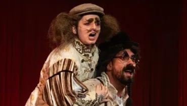 Fotografia em plano médio de dois atores caracterizados em cena teatral cômica e dramática sobre o palco. Em primeiro plano, do centro para a direita, sobressai-se um ator de pele clara, barba escura e trança fina no queixo, usando chapéu preto, óculos redondos e sobrancelhas artificiais espessas e arqueadas. Ele está inclinado para a frente com a boca aberta em expressão de espanto ou esforço físico, segurando as mãos da pessoa que está montada sobre suas costas. No plano intermediário, inclinando-se sobre as costas do primeiro ator, destaca-se a segunda figura teatral, de pele clara, cabelos volumosos e desgrenhados sob uma boina marrom, exibindo sobrancelhas e bigodes falsos pintados em tom marrom. Com a boca bem aberta em expressão de grito ou atuação expressiva, veste um figurino de época trabalhado com bordados, gola alta ornamentada e mangas bufantes listradas em tons de bege e marrom. Ao fundo, o cenário é composto por uma cortina ou cortinado teatral vermelho fechado, sob iluminação cênica focada que realça as expressões faciais e os detalhes dos figurinos dos personagens.