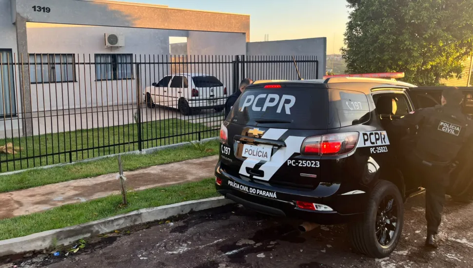 Fotografia em plano geral de uma operação policial realizada pela Polícia Civil do Estado do Paraná (PCPR) em área residencial. Em primeiro plano, à direita, sobressai a traseira de uma viatura caracterizada do modelo SUV de cor preta. No vidro traseiro e na lataria, leem-se as inscrições em letras brancas "PCPR", "POLÍCIA", "POLÍCIA CIVIL DO PARANÁ", o número de emergência "197" e a identificação do veículo "PC-25023". Um policial civil de costas, vestindo jaqueta e calça pretas com o patch "POLÍCIA CIVIL" nas costas, apoia-se junto à porta aberta do passageiro. No plano intermediário, atrás da viatura, observa-se uma grade metálica preta e um portão de ferro que delimitam a frente de uma residência térrea moderna de cor cinza-clara, identificada pelo número "1319". No quintal interno, há um carro branco estacionado e um trecho de gramado. Ao fundo, a paisagem exibe vegetação verde e o horizonte sob iluminação natural suave do amanhecer ou entardecer.
