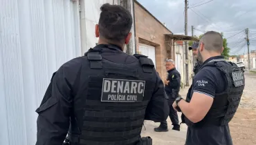 Fotografia em plano médio de policiais civis da Divisão Estadual de Narcóticos (DENARC) da Polícia Civil do Estado do Paraná em ação de campo residencial. Em primeiro plano, no centro e à esquerda, destaca-se a figura de costas de um policial civil de porte físico forte, vestindo jaqueta preta de mangas compridas e colete tático operatório preto no qual se lê o patch sobressalente "DENARC - POLÍCIA CIVIL" em letras brancas. À direita, outro policial civil de pele clara, de perfil, veste camisa polo escura com a inscrição "POLÍCIA CIVIL" no peito e colete tático preto com a sigla "DENARC". No plano intermediário, um terceiro policial civil de cabelo grisalho e óculos de grau, vestindo uniforme escuro e colete de segurança, caminha pelo passeio da rua em direção às casas. Ao fundo, a cena mostra a fachada de residências térreas urbanas com portões brancos e paredes revestidas, ao longo de uma rua sob céu nublado com iluminação natural uniforme do dia.