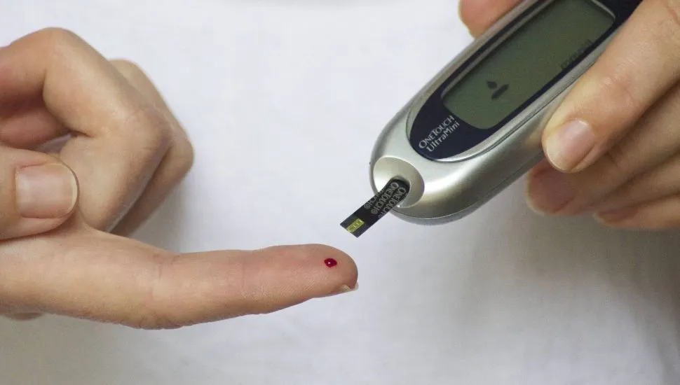 Imagem mostra uma pessoa medindo a glicemia em um aparelho eletrônico. O paciente está com uma gota de sangue no dedo e com o aparelho cinza e preto se aproximando.