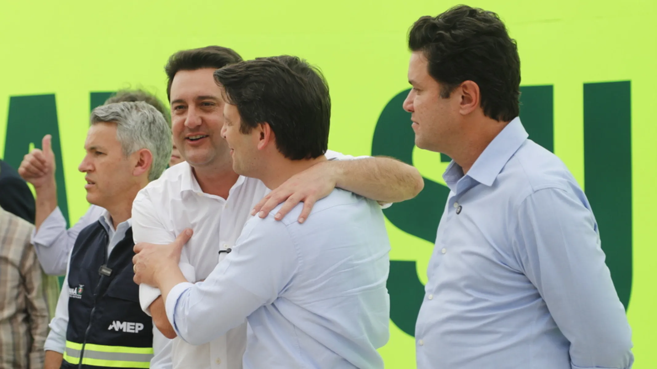 Fotografia em plano médio de autoridades e figuras públicas políticas do Paraná reunidas em evento institucional aberto. Em primeiro plano, do centro para a direita, sobressai-se o governador do Paraná, Ratinho Junior, homem de pele clara e cabelos escuros, vestindo camisa social azul-clara de mangas compridas, posando de perfil com os braços para trás e sorrindo enquanto observa a interação ao lado. Ao seu lado, no centro, dois homens se abraçam calorosamente: um de costas vestindo camisa azul-clara envolve o ombro de Ratinho Junior, e o outro, de pele clara e camisa branca, sorri de frente retribuindo o abraço. À esquerda, em primeiro plano, outro integrante de pele clara e cabelo grisalho veste colete tático escuro de serviço público com a sigla institucional "AMEP" e faixa refletiva amarela. Ao fundo, à extrema esquerda, observa-se uma pessoa fazendo o sinal de "joia" com o polegar erguido. No plano intermediário e ao fundo, o cenário é composto por um grande painel publicitário ou institucional em tom verde-limão com letras grandes em verde-escuro, sob iluminação natural do dia.