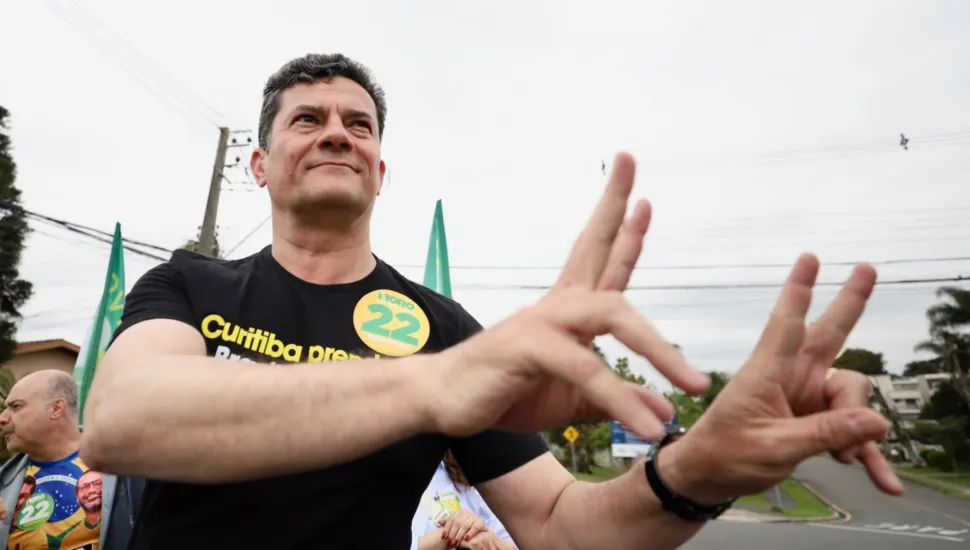 Fotografia em plano médio de autoridades e figuras públicas políticas do Paraná reunidas em evento institucional aberto. Em primeiro plano, do centro para a direita, sobressai-se o governador do Paraná, Ratinho Junior, homem de pele clara e cabelos escuros, vestindo camisa social azul-clara de mangas compridas, posando de perfil com os braços para trás e sorrindo enquanto observa a interação ao lado. Ao seu lado, no centro, dois homens se abraçam calorosamente: um de costas vestindo camisa azul-clara envolve o ombro de Ratinho Junior, e o outro, de pele clara e camisa branca, sorri de frente retribuindo o abraço. À esquerda, em primeiro plano, outro integrante de pele clara e cabelo grisalho veste colete tático escuro de serviço público com a sigla institucional "AMEP" e faixa refletiva amarela. Ao fundo, à extrema esquerda, observa-se uma pessoa fazendo o sinal de "joia" com o polegar erguido. No plano intermediário e ao fundo, o cenário é composto por um grande painel publicitário ou institucional em tom verde-limão com letras grandes em verde-escuro, sob iluminação natural do dia.