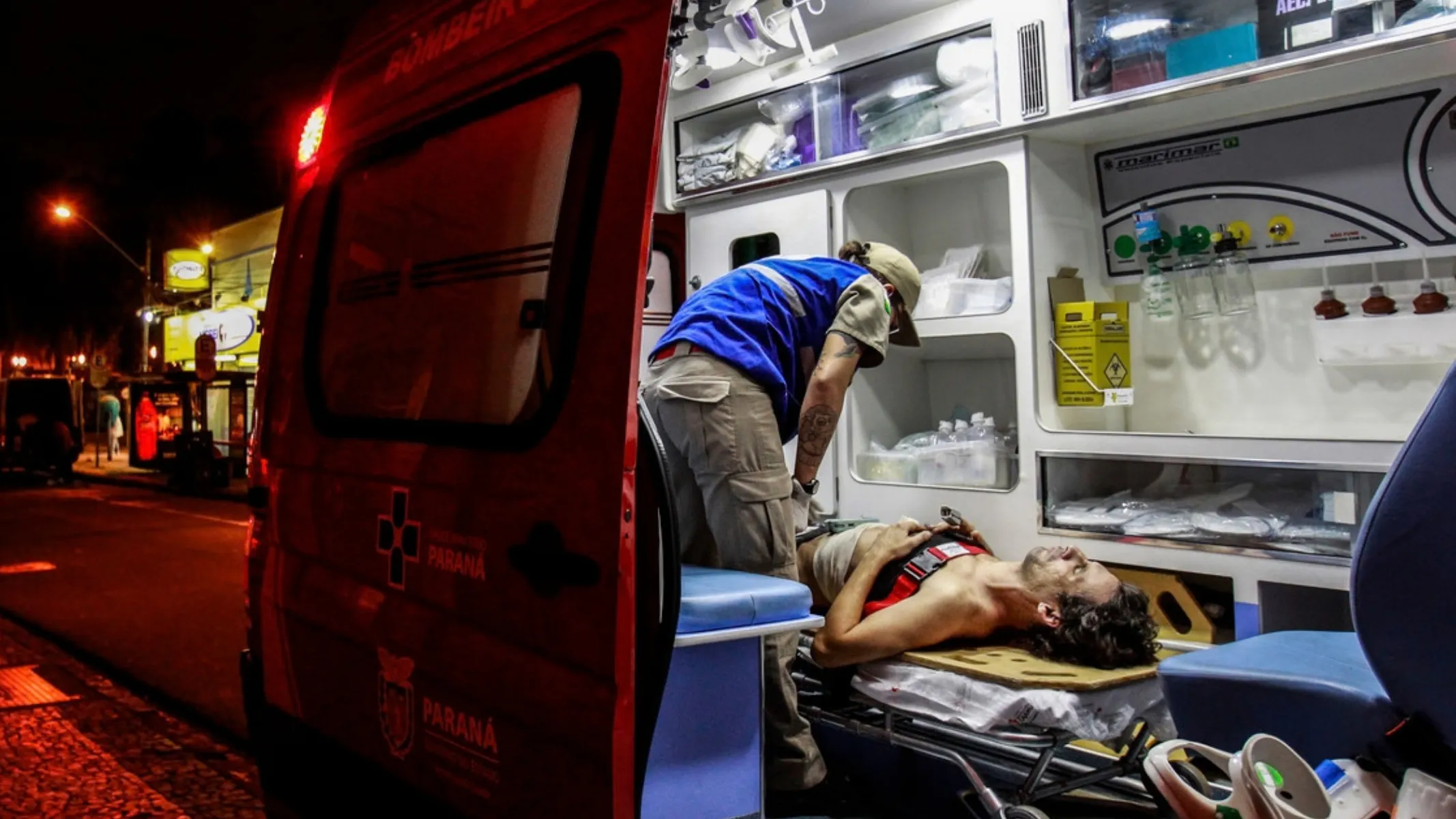 Fotografia em plano médio do interior de uma ambulância de resgate durante atendimento pré-hospitalar noturno. Em primeiro plano, à esquerda, destaca-se a porta traseira aberta da ambulância de cor vermelha, onde sobressaem a luz vermelha do giroflex aceso e as inscrições institucionais "BOMBEIROS" no topo e "PARANÁ GOVERNO DO ESTADO" com a cruz do resgate na lataria. No plano intermediário, no interior bem iluminado do veículo de socorro, um homem ferido e sem camisa jaz deitado sobre a maca dobrável com o tórax enfaixado e tiras de fixação de emergência. Curvado sobre ele, um socorrista de boné bege, colete azul e calça cargo caqui presta os primeiros socorros e manipula equipamentos médicos. A estrutura interna da ambulância exibe prateleiras brancas organizadas com insumos hospitalares, frascos de soro, caixas descartadoras de perfurocortantes e cilindros de oxigênio. Ao fundo, à esquerda, vê-se a calçada de petit-pavé e a iluminação pública amarelada de uma rua urbana durante a noite.