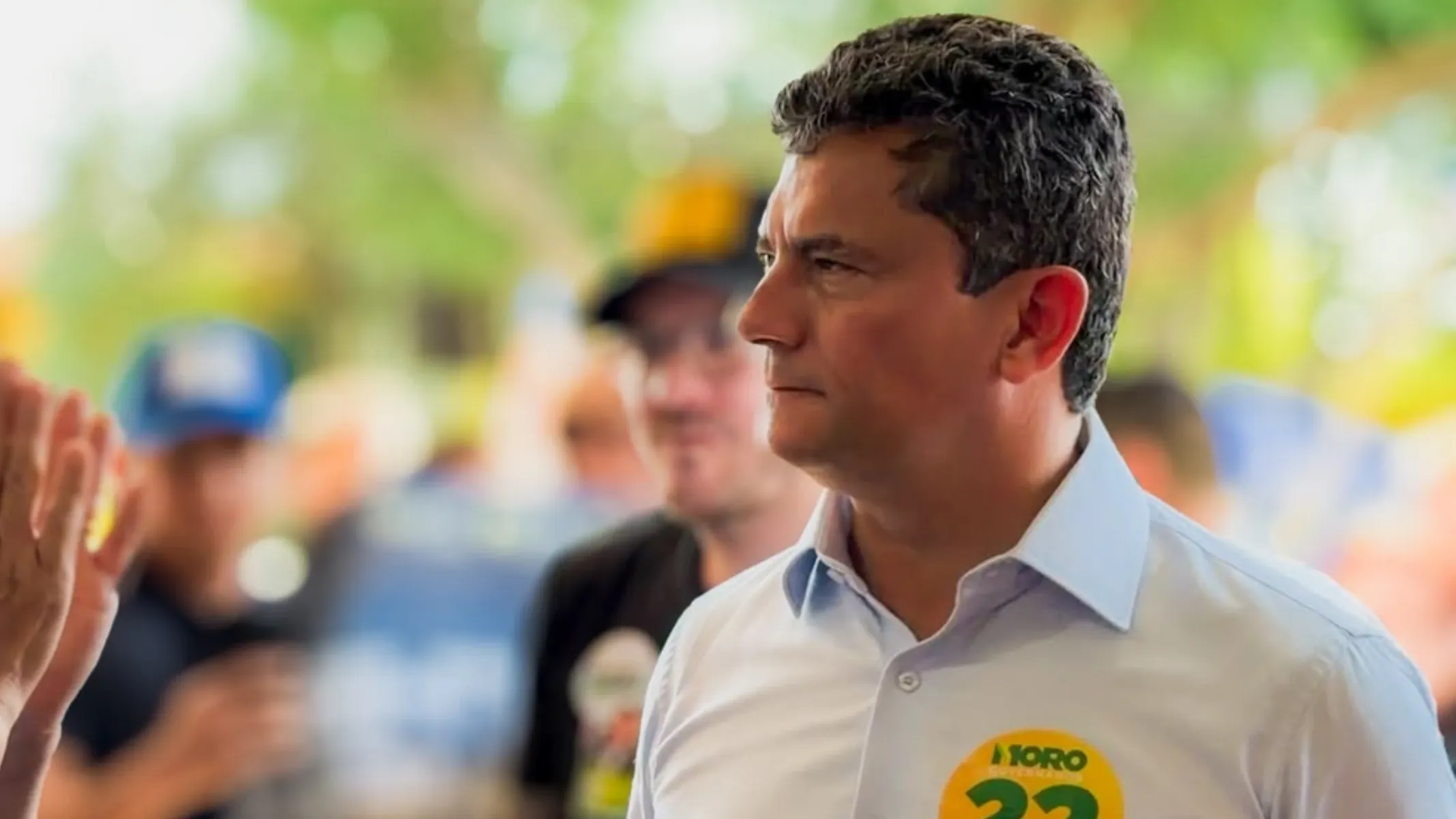 Fotografia em plano médio do político Sergio Moro durante evento público de campanha eleitoral ao ar livre. Em primeiro plano, à direita, destaca-se o ex-juiz e político Sergio Moro, homem de pele clara e cabelos curtos escuros levemente acinzentados nas fontes. Visto em semi-perfil voltado para a esquerda e enquadrado do peito para cima, ele veste camisa social de mangas compridas em tom azul-claro. No peito, sobressai um adesivo circular amarelo de campanha com o nome "MORO" e o número "22" impresso em destaque. À extrema esquerda, observa-se em primeiro plano a mão espalmada de um popular cumprimentando a figura pública. No plano intermediário e ao fundo, desfocados pela reduzida profundidade de campo, leem-se rostos e figuras de apoiadores e integrantes da equipe, alguns vestindo bonés e camisetas escuras com adesivos de campanha. Ao fundo, a iluminação natural diurna filtra-se através da copa de árvores e vegetação verde.