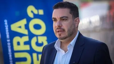 Paulo Martins olha pa ao horizonta, em frente a um banner. ele veste um paletó azul e camisa clara, sem gravata