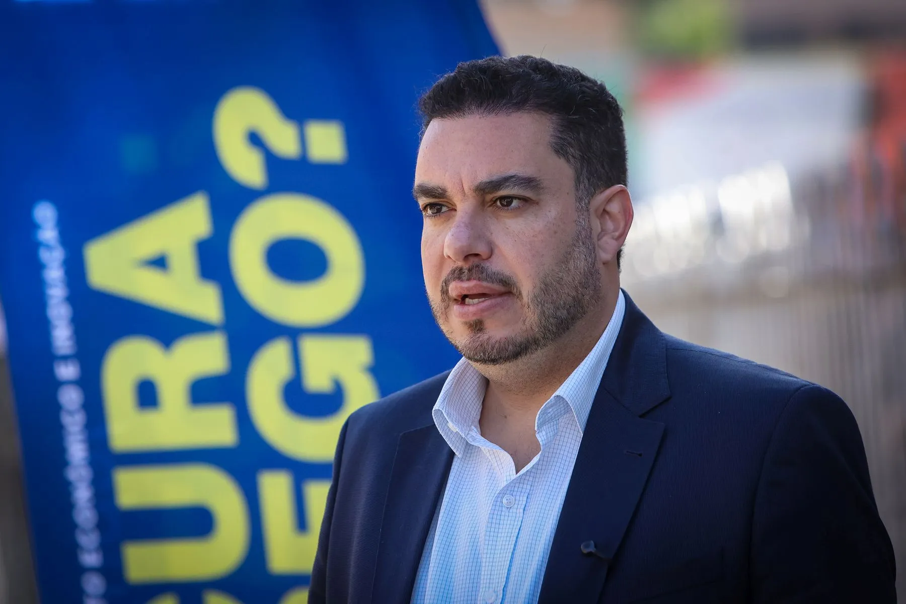 Paulo Martins olha pa ao horizonta, em frente a um banner. ele veste um paletó azul e camisa clara, sem gravata