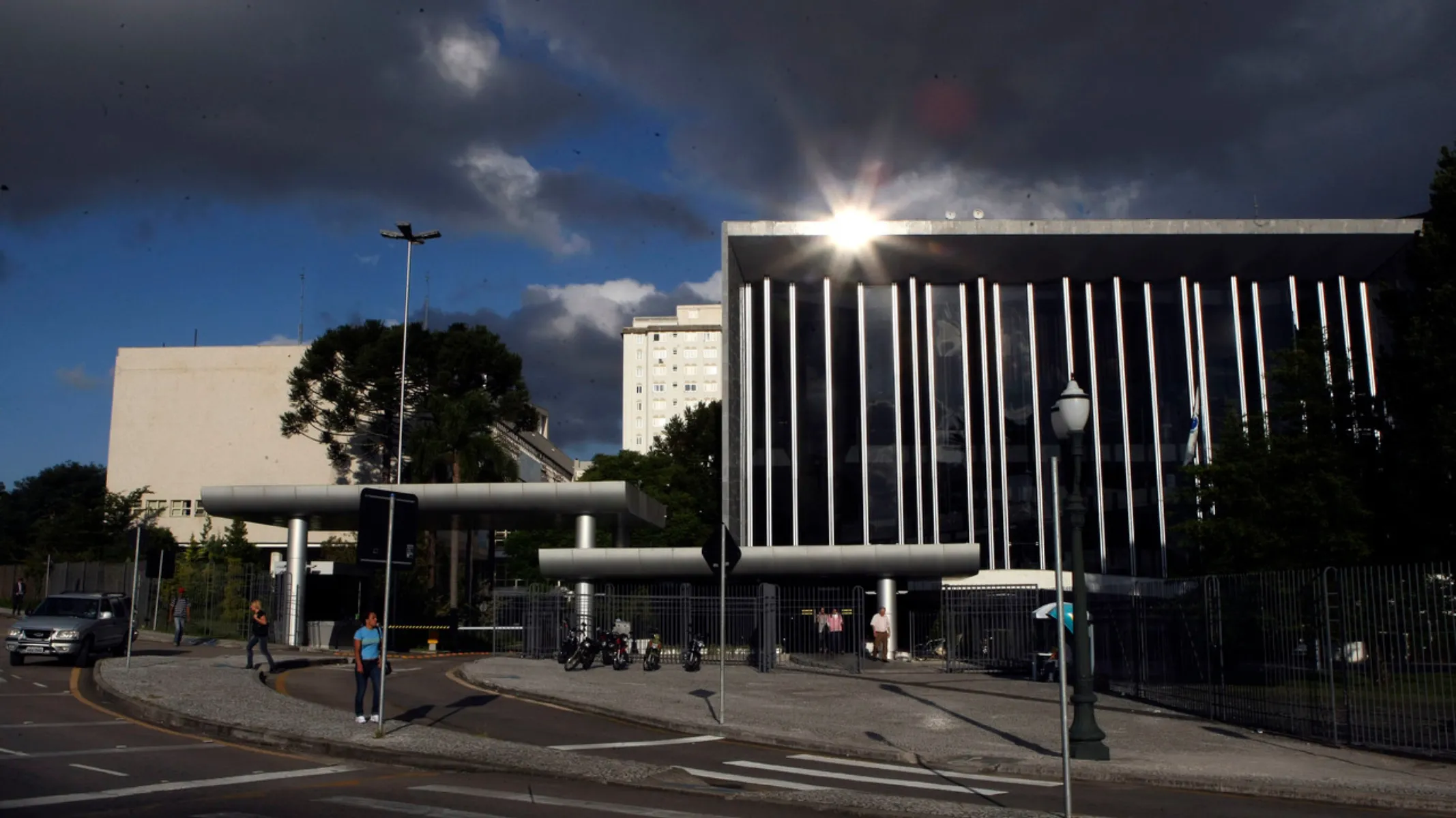 Fotografia em plano geral da fachada do Palácio Iguaçu, sede do Poder Executivo do Estado do Paraná, localizado no Centro Cívico em Curitiba. Em primeiro plano, observa-se a calçada de acesso, a faixa de pedestres no asfalto e a via de trânsito em curva. À esquerda, pedestres transitam pelo passeio e um veículo prata trafega pela pista. À direita, destaca-se um poste de iluminação pública em estilo clássico e um gradil metálico de proteção no perímetro do prédio. No plano intermediário, estende-se a fachada moderna do Palácio Iguaçu, caracterizada por uma ampla colunata vertical de concreto e grandes painéis de vidro espelhado. Um reflexo solar intenso (efeito starburst) incide sobre o topo superior esquerdo da cobertura metálica do edifício. Abaixo e à esquerda, veem-se as estruturas de portaria e marquises de acesso ao complexo governamental, com motocicletas e pedestres próximos aos portões. Ao fundo, sobressaem-se edifícios residenciais e administrativos da região central, copas de árvores nativas — incluindo uma araucária — e o céu com nuvens escuras e densas sob iluminação natural dramática de fim de tarde.