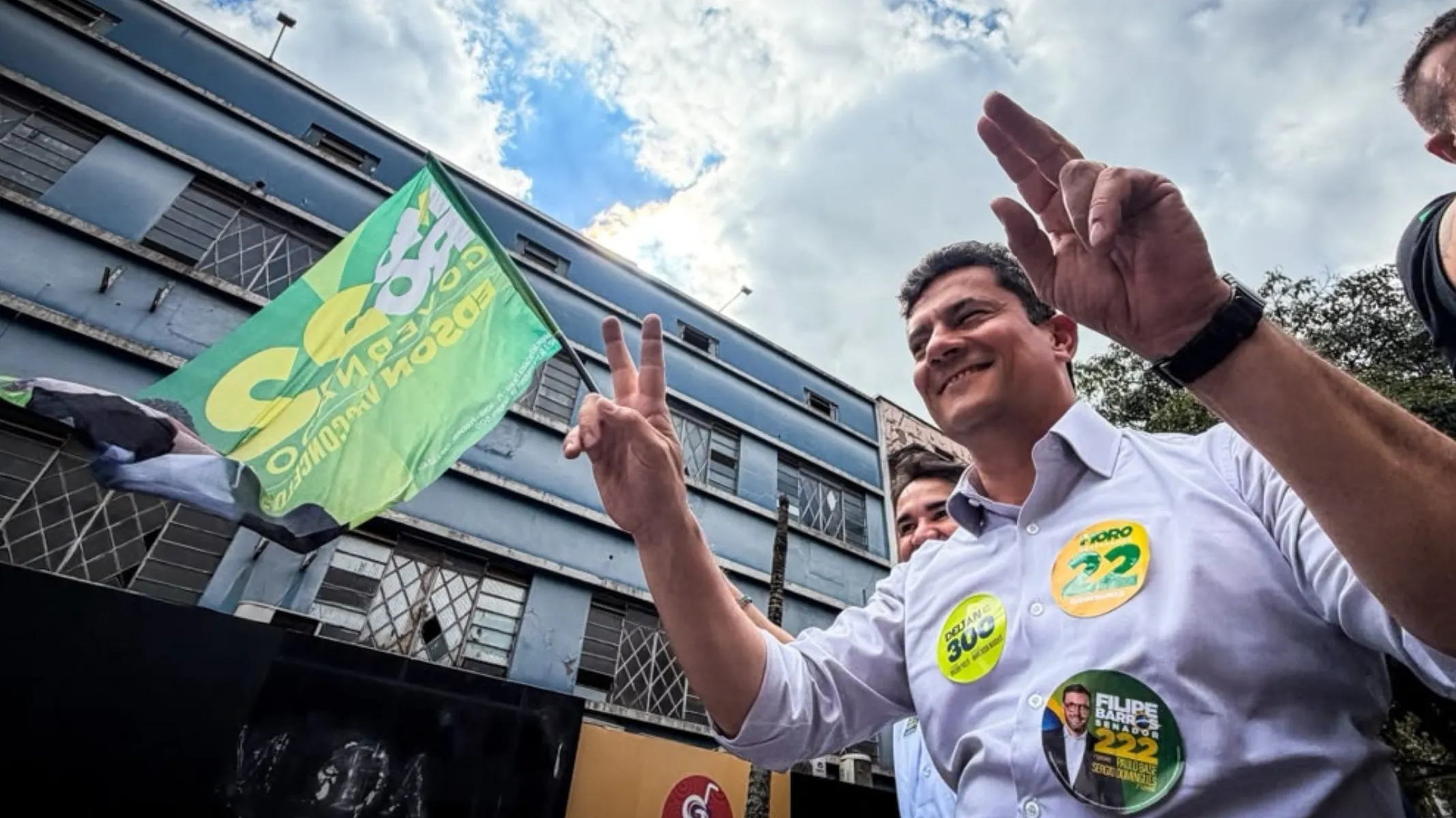 Fotografia em contra-plongée (ângulo de baixo para cima) em plano médio do político Sergio Moro participando de ato público de campanha eleitoral em rua urbana. Em primeiro plano, à direita e no centro, destaca-se Sergio Moro, homem de pele clara e cabelos curtos escuros, sorrindo e fazendo o sinal de vitória ("V") com os dedos indicador e médio levantados em ambas as mãos. Ele veste camisa social branca de mangas compridas dobradas até os antebraços. No peito da camisa, sobressaem adesivos circulares de campanha: um amarelo com o nome "MORO" e o número "19", outro amarelo-limão com a inscrição "DELTAN 300" e um terceiro adesivo de candidato a deputado com a imagem de Filipe Barros. À esquerda, em primeiro plano, uma mão segura uma bandeira de campanha verde e amarela em movimento. No plano intermediário, atrás de Moro, observa-se parcialmente o rosto de outro integrante da equipe acompanhando a caminhada. Ao fundo, a arquitetura urbana exibe a fachada de um prédio comercial de tom azul com janelas retangulares sob céu com nuvens e iluminação natural diurna.