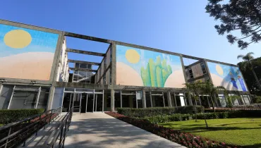 Fotografia em plano geral da fachada do Palácio Iguaçu, sede do Poder Executivo do Estado do Paraná, localizado no Centro Cívico em Curitiba. Em primeiro plano, observa-se uma rampa de acesso pavimentada para pedestres e pessoas com mobilidade reduzida, guarnecida por corrimãos metálicos pretos. À direita da rampa, estendem-se jardins bem cuidados com gramado verde, canteiros de flores avermelhadas e arbustos podados. No plano intermediário, destaca-se a imponente fachada de concreto do edifício governamental, marcada por grandes painéis artísticos de azulejos coloridos que retratam elementos paisagísticos e sóis estilizados. A entrada principal possui amplas portas e janelas de vidro emolduradas por estruturas metálicas. Ao fundo, à direita, sobressaem-se os galhos de uma árvore de araucária contra o céu limpo e azul sob forte luz solar diurna.