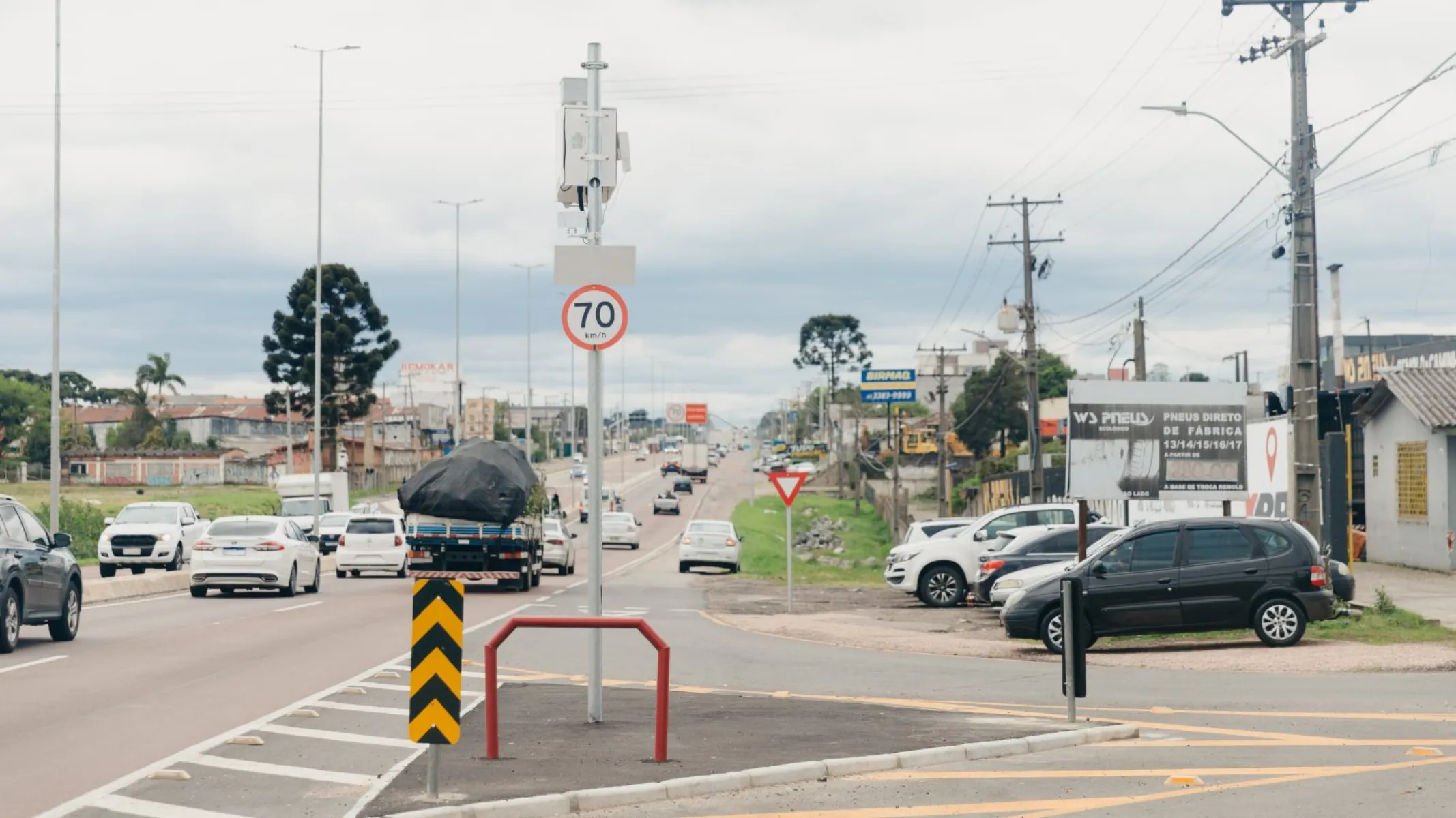 Fotografia em plano geral de uma rodovia asfaltada movimentada em área urbana ou metropolitana, sob céu nublado e iluminação natural difusa. Em primeiro plano, no centro, destaca-se uma placa de sinalização de velocidade máxima permitida indicando "70 km/h", afixada em um poste metálico que também suporta equipamentos eletrônicos de fiscalização de trânsito. Abaixo da placa, há um delineador de curva com setas pretas e amarelas e uma estrutura metálica de proteção vermelha na ilha divisora da pista. À direita, veículos encontram-se estacionados em um recuo próximo a um estabelecimento comercial, onde se lê uma placa publicitária de pneus. No plano intermediário, à esquerda, o fluxo de carros e utilitários trafega no sentido oposto ou na mesma direção da via pavimentada, ladeada por sinalizações horizontais e margens com vegetação rasteira. Ao fundo, a rodovia estende-se em perspectiva ao longo da paisagem urbana, com postes de rede elétrica, árvores — incluindo araucárias — e construções ao longo das margens sob um horizonte de céu nublado.