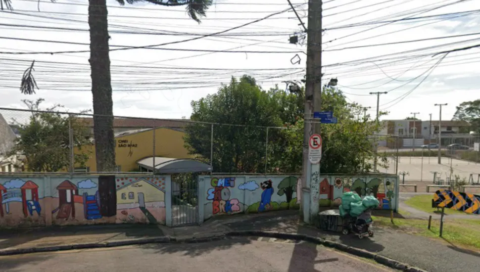 Fotografia em plano geral da fachada do Centro Municipal de Educação Infantil (CMEI) São Brás, situado em uma esquina urbana no bairro São Brás, em Curitiba. Em primeiro plano, observa-se a calçada de paralelepípedos na esquina, delimitada por um muro mureta de alvenaria pintado com murais coloridos e ilustrações infantis que retratam parquinhos e a identificação "CMEI SÃO BRÁS". Um poste de energia elétrica de concreto ergue-se próximo à esquina, ostentando uma placa de trânsito de proibido estacionar e placas de identificação de rua. Na base do poste, há um carrinho de carga com sacolas e volumes. No plano intermediário, atrás da mureta, estende-se uma cerca de tela metálica e o prédio amarelo do CMEI, parcialmente sombreado pelo tronco de uma grande araucária à esquerda e por árvores e vegetação no entorno. À direita, avista-se uma praça ou quadra esportiva aberta com traves e alambrado. Ao fundo, a paisagem urbana exibe o cruzamento de ruas, casas residenciais e um denotado emaranhado de cabos e fios de rede elétrica cruzando o céu sob iluminação natural diurna.