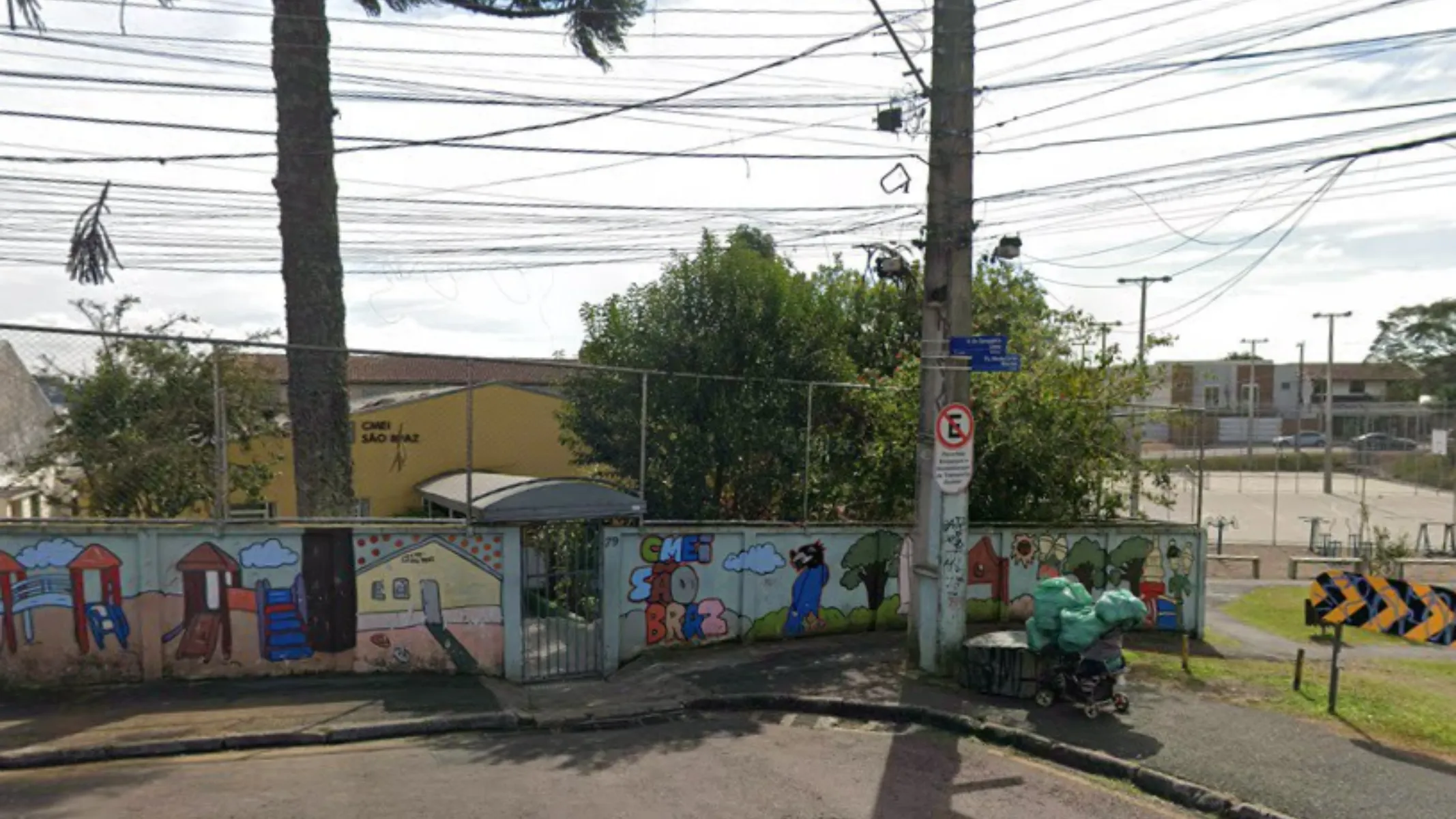 Fotografia em plano geral da fachada do Centro Municipal de Educação Infantil (CMEI) São Brás, situado em uma esquina urbana no bairro São Brás, em Curitiba. Em primeiro plano, observa-se a calçada de paralelepípedos na esquina, delimitada por um muro mureta de alvenaria pintado com murais coloridos e ilustrações infantis que retratam parquinhos e a identificação "CMEI SÃO BRÁS". Um poste de energia elétrica de concreto ergue-se próximo à esquina, ostentando uma placa de trânsito de proibido estacionar e placas de identificação de rua. Na base do poste, há um carrinho de carga com sacolas e volumes. No plano intermediário, atrás da mureta, estende-se uma cerca de tela metálica e o prédio amarelo do CMEI, parcialmente sombreado pelo tronco de uma grande araucária à esquerda e por árvores e vegetação no entorno. À direita, avista-se uma praça ou quadra esportiva aberta com traves e alambrado. Ao fundo, a paisagem urbana exibe o cruzamento de ruas, casas residenciais e um denotado emaranhado de cabos e fios de rede elétrica cruzando o céu sob iluminação natural diurna.