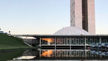 Fotografia em plano geral do Congresso Nacional do Brasil, localizado em Brasília, captado ao entardecer. Em primeiro plano, estende-se o espelho d'água frontal do complexo arquitetônico, refletindo com clareza a fachada do edifício e o tom alaranjado do céu do crepúsculo. À esquerda, vê-se um talude inclinado com gramado verde, margeado por uma via elevada onde automóveis estão estacionados ou em circulação. À direita, veículos estacionados alinham-se ao longo da esplanada. No plano intermediário, destaca-se a arquitetura modernista de Oscar Niemeyer: a base horizontal com janelas retangulares enquadradas por colunas e a cúpula convexa voltada para cima da Câmara dos Deputados. Ao fundo, ergue-se uma das imensas torres gêmeas verticais do Congresso Nacional, com revestimento em mármore claro e janelas em venezianas verticais, sob um céu limpo de fim de tarde.