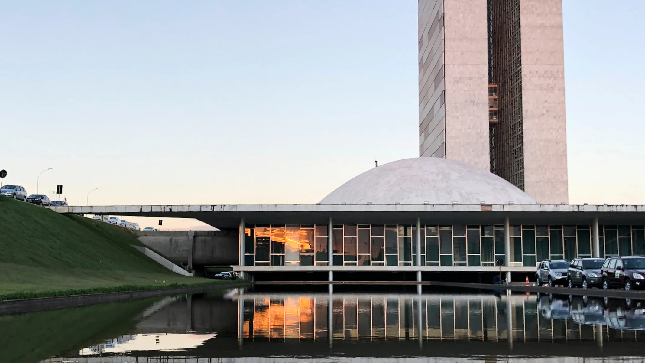 Fotografia em plano geral do Congresso Nacional do Brasil, localizado em Brasília, captado ao entardecer. Em primeiro plano, estende-se o espelho d'água frontal do complexo arquitetônico, refletindo com clareza a fachada do edifício e o tom alaranjado do céu do crepúsculo. À esquerda, vê-se um talude inclinado com gramado verde, margeado por uma via elevada onde automóveis estão estacionados ou em circulação. À direita, veículos estacionados alinham-se ao longo da esplanada. No plano intermediário, destaca-se a arquitetura modernista de Oscar Niemeyer: a base horizontal com janelas retangulares enquadradas por colunas e a cúpula convexa voltada para cima da Câmara dos Deputados. Ao fundo, ergue-se uma das imensas torres gêmeas verticais do Congresso Nacional, com revestimento em mármore claro e janelas em venezianas verticais, sob um céu limpo de fim de tarde.