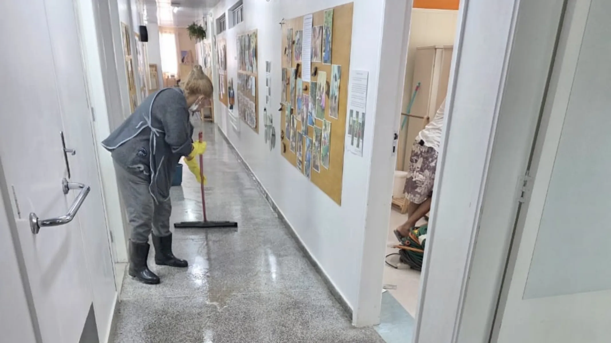 Fotografia em plano médio do corredor interno de uma escola ou instituição de ensino, mostrando uma atividade de limpeza e manutenção. Em primeiro plano, à esquerda, uma trabalhadora de limpeza com cabelos loiros presos, vestindo blusa e calça cinzas, avental, luvas amarelas de borracha e botas pretas de cano alto, empunha um rodo de cabo vermelho para secar o piso de granito molhado. À direita, pela abertura de uma porta, vê-se parcialmente outra pessoa no interior de uma sala organizando materiais e equipamentos. No plano intermediário, as paredes brancas do corredor exibem murais e cartazes informativos com fotografias e trabalhos pedagógicos fixados com tachinhas. Ao fundo, o corredor estende-se em perspectiva, iluminado por luz natural difusa proveniente de janelas ao longe.