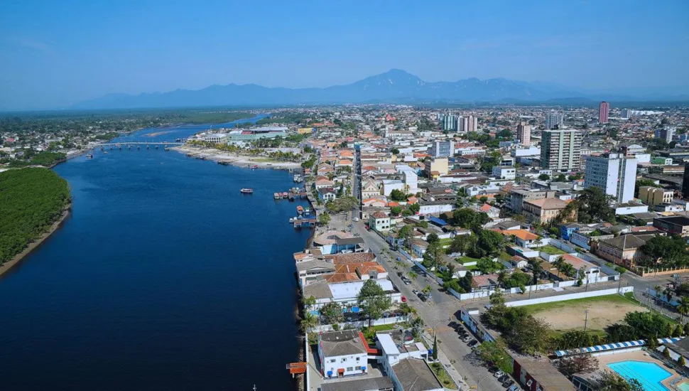 Fotografia aérea em plano geral da cidade de Antonina, no litoral do Paraná, banhada pelas águas da Baía de Antonina. Em primeiro plano e à esquerda, estende-se a extensa faixa de águas escuras da baía, margeada por áreas de manguezal verdejante. À direita, avista-se a malha urbana da cidade, com ruas arborizadas, construções históricas, residências com telhados coloniais e alguns edifícios mais altos. No plano intermediário, a orla urbana acompanha o contorno da baía, onde se destacam pequenas embarcações atracadas e ruas que serpenteiam a paisagem litorânea. Ao fundo, sobressaem-se imponentes cadeias de montanhas e serras cobertas pela Mata Atlântica sob um céu azul claro e limpo, iluminado pela luz solar diurna.