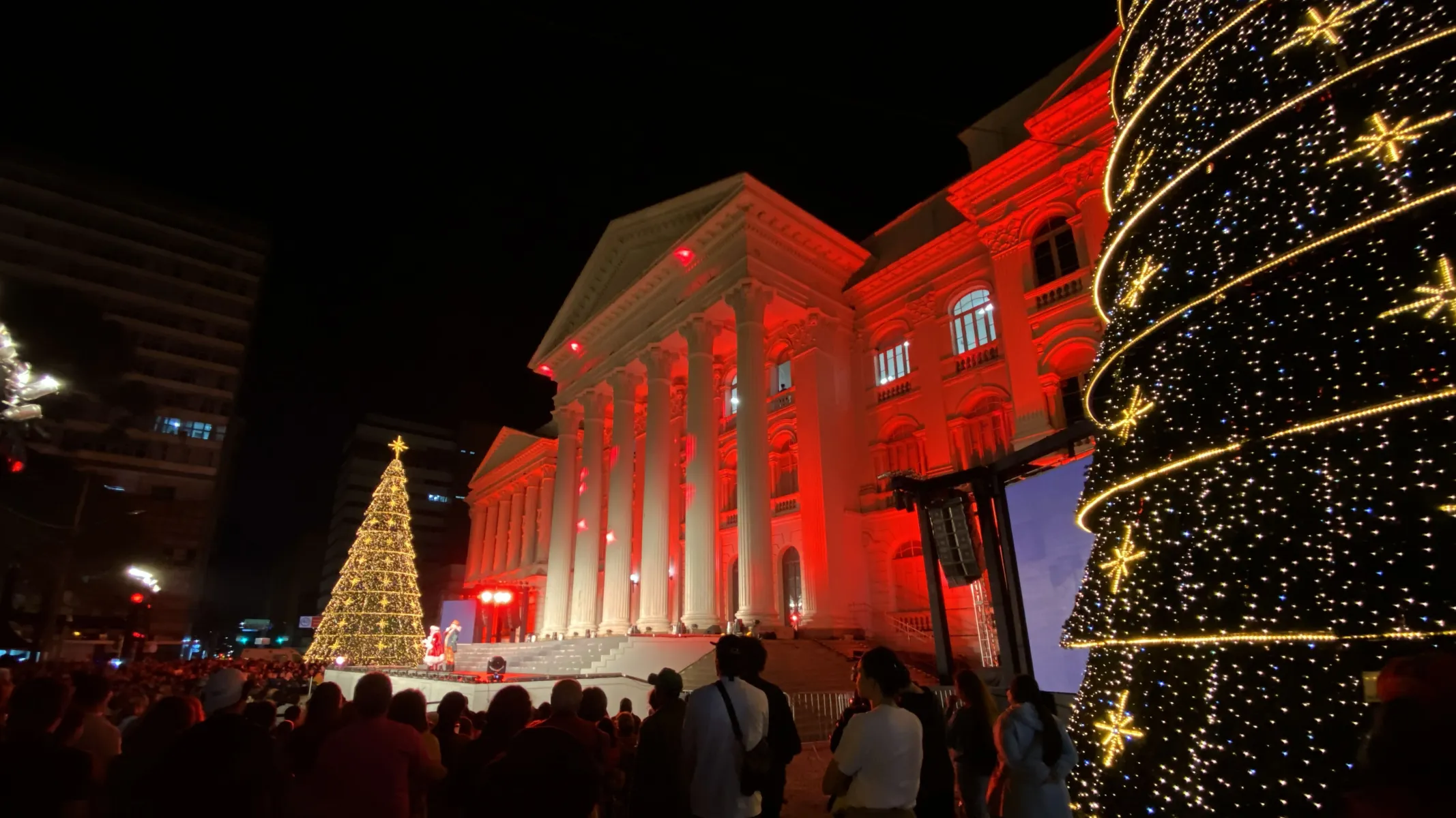Fotografia em plano geral de um evento noturno festivo de Natal em praça pública, tendo ao fundo a fachada iluminada de um edifício histórico, semelhante à Universidade Federal do Paraná (UFPR) na Praça Santos Andrade. Em primeiro plano e à direita, destaca-se a estrutura parcial de uma imensa árvore de Natal troncônica ornamentada com milhares de pequenas luzes douradas e enfeites em formato de estrela. Na parte inferior, uma multidão de pessoas de costas assiste à celebração. No plano intermediário, o imponente edifício neoclássico exibe colunas imponentes banhadas por uma intensa iluminação cênica vermelha. À esquerda, avista-se uma segunda árvore de Natal iluminada e figuras decorativas no palco montado em frente à fachada. Ao fundo, a escuridão da noite contrasta com a forte iluminação cenográfica natalina e as luzes de edifícios ao redor.