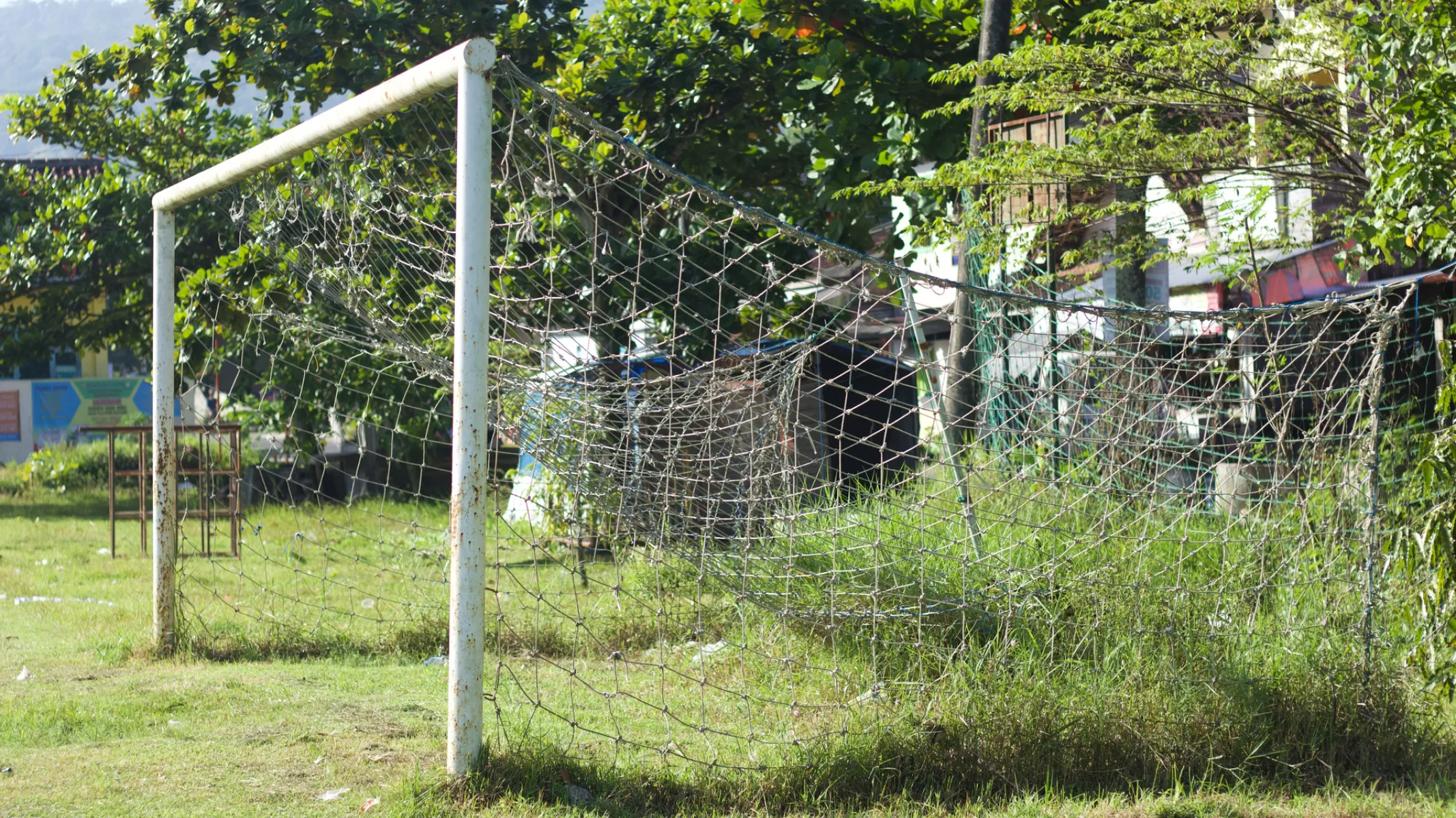 Fotografia em plano médio de uma trave de futebol desgastada, instalada em um campo de grama irregular. Em primeiro plano, destaca-se a estrutura tubular metálica branca da trave, apresentando sinais de oxidação e desgaste pelo tempo. A rede de corda branca está danificada, com rasgos e áreas frouxas onde a grama alta e mato crescem por entre as malhas. No plano intermediário, o campo apresenta vegetação rasteira irregular e pequenos arbustos ao redor. Ao fundo, árvores frondosas e construções residenciais compõem o cenário sob a luz natural diurna.