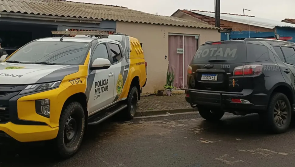 Fotografia em plano médio de duas viaturas policiais estacionadas em uma rua residencial pavimentada, diante da fachada de uma residência. Em primeiro plano e à esquerda, destaca-se uma caminhonete da Polícia Militar do Paraná (3º BPM), com pintura caracterizada em branco e amarelo, ostentando o brasão oficial e a inscrição "POLÍCIA MILITAR" na lateral. À direita, posiciona-se uma viatura SUV preta da ROTAM, exibindo a identificação tática "ROTAM" no vidro traseiro e na carroceria, além da placa visível e numeração operacional. No plano intermediário, as viaturas ocupam a via em frente a uma casa térrea de alvenaria com telhado de telhas de barro e uma porta de correr de vidro. Ao fundo, sob uma luz natural difusa de dia nublado, o cenário urbano residencial compõe o ambiente da operação de segurança.