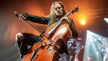 Fotografia em plano contrapilado de um violoncelista performando energeticamente durante um show ao vivo, sob iluminação cênica dramática. Em primeiro plano, destaca-se o instrumento de madeira envernizada e o arco em riste sendo empunhado com intensidade pelo músico. O artista, um homem de cabelos longos e claros, barba e expressão concentrada, veste roupas pretas, incluindo uma jaqueta e calças escuras. Ele adota uma postura dinâmica no palco. No plano intermediário, feixes de luz branca e quente cortam a penumbra ao fundo, realçando a fumaça de palco e a atmosfera do concerto. Ao fundo, estruturas metálicas de iluminação e painéis completam o cenário da apresentação musical.