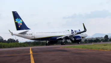 Fotografia em plano geral de uma aeronave comercial da companhia aérea Azul taxiando em uma pista de aeroporto. Em primeiro plano, a pista de pouso e decolagem exibe pavimentação asfáltica escura com sinalizações horizontais pintadas em amarelo. No plano intermediário, o avião comercial de fuselagem branca e detalhes em azul e amarelo desloca-se pela pista. A cauda da aeronave exibe a pintura estilizada com o mapa do Brasil composto por blocos coloridos, além da matrícula "PR-YRZ" visível na fuselagem traseira. Os motores turbofan encontram-se sob as asas e o trem de aterrissagem está apoiado no solo. Ao fundo, a linha do horizonte revela uma área verde com vegetação arbórea e colinas sob um céu parcialmente nublado com luz natural diurna.