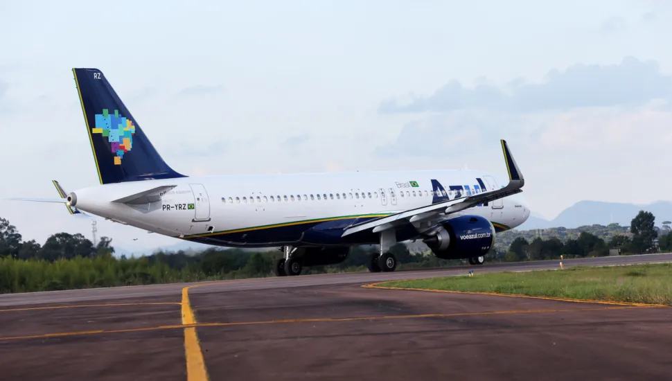 Fotografia em plano geral de uma aeronave comercial da companhia aérea Azul taxiando em uma pista de aeroporto. Em primeiro plano, a pista de pouso e decolagem exibe pavimentação asfáltica escura com sinalizações horizontais pintadas em amarelo. No plano intermediário, o avião comercial de fuselagem branca e detalhes em azul e amarelo desloca-se pela pista. A cauda da aeronave exibe a pintura estilizada com o mapa do Brasil composto por blocos coloridos, além da matrícula "PR-YRZ" visível na fuselagem traseira. Os motores turbofan encontram-se sob as asas e o trem de aterrissagem está apoiado no solo. Ao fundo, a linha do horizonte revela uma área verde com vegetação arbórea e colinas sob um céu parcialmente nublado com luz natural diurna.