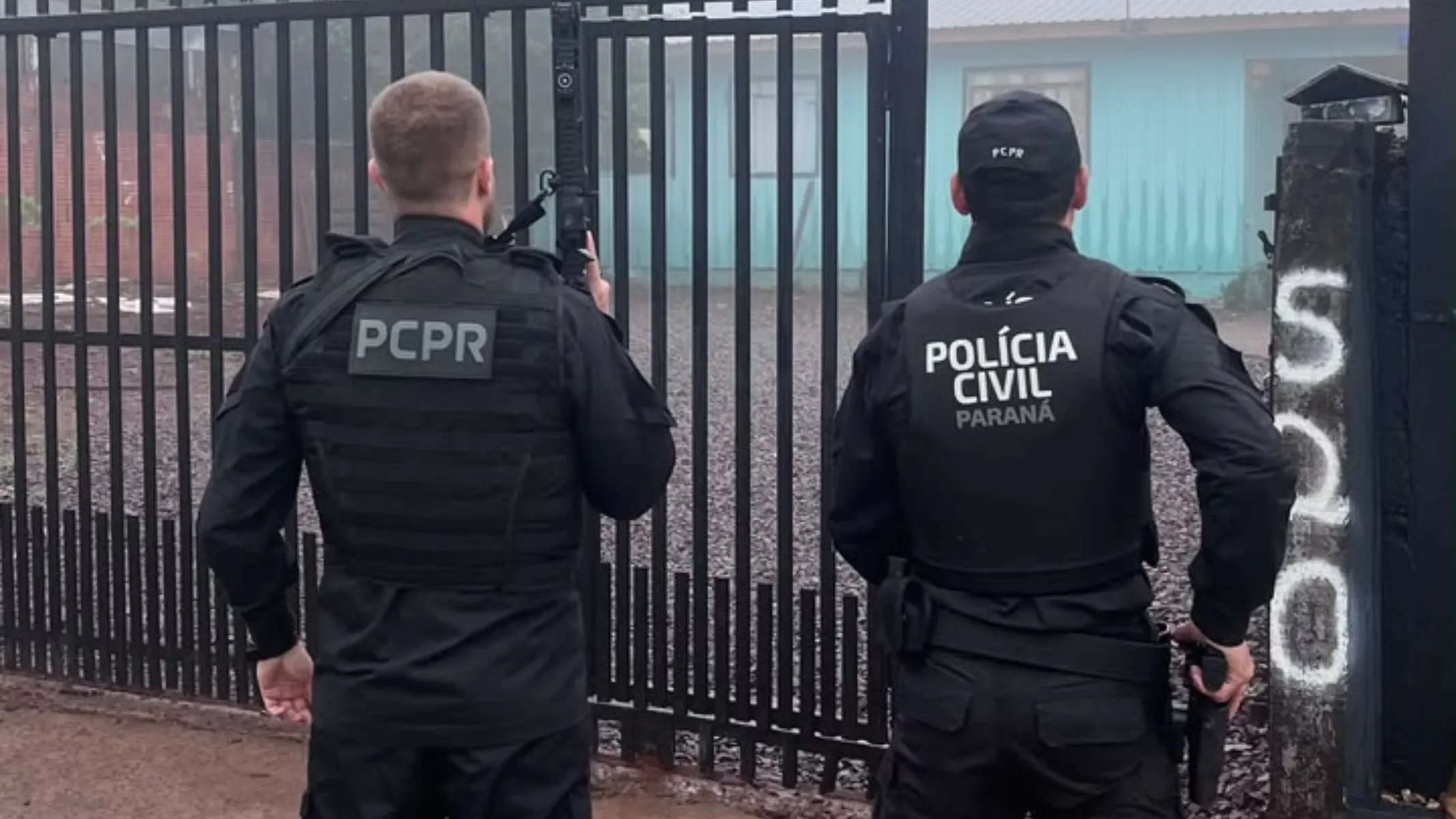 Fotografia em plano médio de costas de dois policiais civis paranaenses durante uma operação de segurança em frente a um imóvel. Em primeiro plano e à esquerda, um policial civil veste uniforme preto composto por camisa de manga longa, calça e um colete tático equipado com um patch cinza com a sigla "PCPR". Ele segura um fuzil verticalmente junto ao corpo. À direita, o segundo policial civil usa vestimenta preta tática similar, boné escuro com a inscrição "PCPR", e um colete com o brasão e os dizeres "POLÍCIA CIVIL PARANÁ". Ele porta coldre e equipamento na cintura. No plano intermediário, os agentes estão posicionados de costas, diante de um portão metálico preto de grades verticais. Ao lado direito do portão, um pilar de concreto exibe o número "520" pintado em branco na vertical. Ao fundo, avista-se a fachada de uma residência com paredes em tom azulado sob a luz difusa do dia.