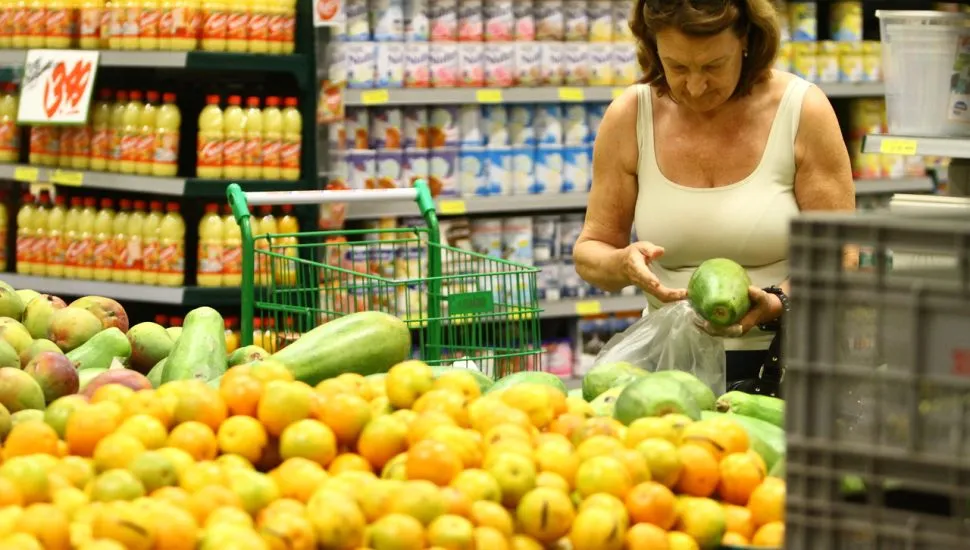 Fotografia em plano médio de uma mulher fazendo compras no setor de hortifrúti de um supermercado. Em primeiro plano e à esquerda, uma grande pilha de laranjas e frutas cítricas amareladas transborda de caixas expositoras. Ao centro, um carrinho de compras verde repousa entre as bancas. No plano intermediário e à direita, uma mulher de cabelos castanhos curtos e óculos apoiados na cabeça examina uma fruta verde que segura com as duas mãos, colocando-a dentro de um saco plástico transparente. Ela veste regata branca. Ao fundo, prateleiras metálicas repletas de produtos industrializados, como garrafas de suco e enlatados, estendem-se por todo o corredor sob a iluminação interna do estabelecimento.