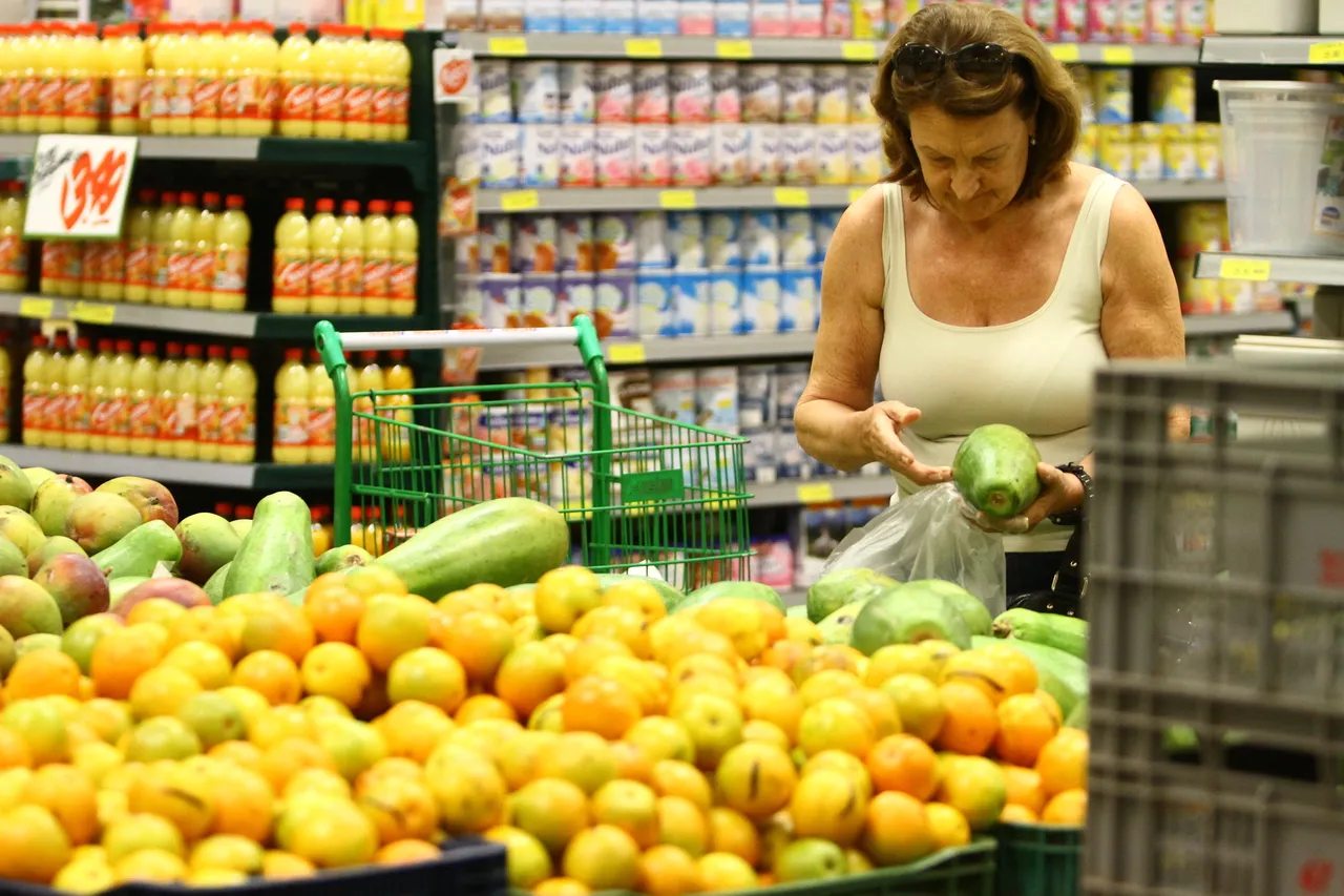 Fotografia em plano médio de uma mulher fazendo compras no setor de hortifrúti de um supermercado. Em primeiro plano e à esquerda, uma grande pilha de laranjas e frutas cítricas amareladas transborda de caixas expositoras. Ao centro, um carrinho de compras verde repousa entre as bancas. No plano intermediário e à direita, uma mulher de cabelos castanhos curtos e óculos apoiados na cabeça examina uma fruta verde que segura com as duas mãos, colocando-a dentro de um saco plástico transparente. Ela veste regata branca. Ao fundo, prateleiras metálicas repletas de produtos industrializados, como garrafas de suco e enlatados, estendem-se por todo o corredor sob a iluminação interna do estabelecimento.
