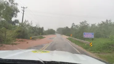 Fotografia em primeiro plano (ponto de vista do motorista) de uma rodovia sob chuva intensa, mostrando uma ponte com acúmulo de lama e água barrenta na pista. Em primeiro plano, o capô branco de um veículo oficial exibe o brasão institucional e as palhetas do limpador de para-brisa acionadas. No plano intermediário, a estrada pavimentada exibe faixas amarelas e divisores metálicos de proteção (guard-rails) nas margens laterais. À direita, uma placa azul sinaliza a "Ponte sobre o Rio Ribeirão Bandeirantes do Sul - Extensão 65 m". A pista e os acostamentos encontram-se cobertos por uma camada de lama avermelhada decorrente de escoamento pluvial. Ao fundo, sob um céu totalmente encoberto e cinzento característico de temporal, a vegetação ribeirinha e os postes de rede elétrica estendem-se ao longo da paisagem chuvosa.