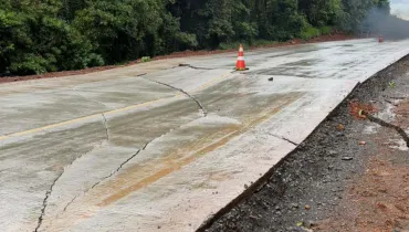 Fotografia em plano médio de uma rodovia pavimentada em concreto que apresenta rachaduras e danos estruturais na pista. Em primeiro plano, a laje de concreto da estrada exibe grandes fissuras transversais e longitudinais que atravessam a faixa de rolamento, sob uma superfície molhada. Um cone de sinalização laranja e branco encontra-se posicionado sobre a pista danificada para alertar motoristas. No plano intermediário, a rodovia estende-se em curva suave, ladeada por encostas de terra vermelha e vegetação densa. Ao fundo, sob um dia nublado e chuvoso, árvores e arbustos compõem a paisagem marginal da estrada afetada.